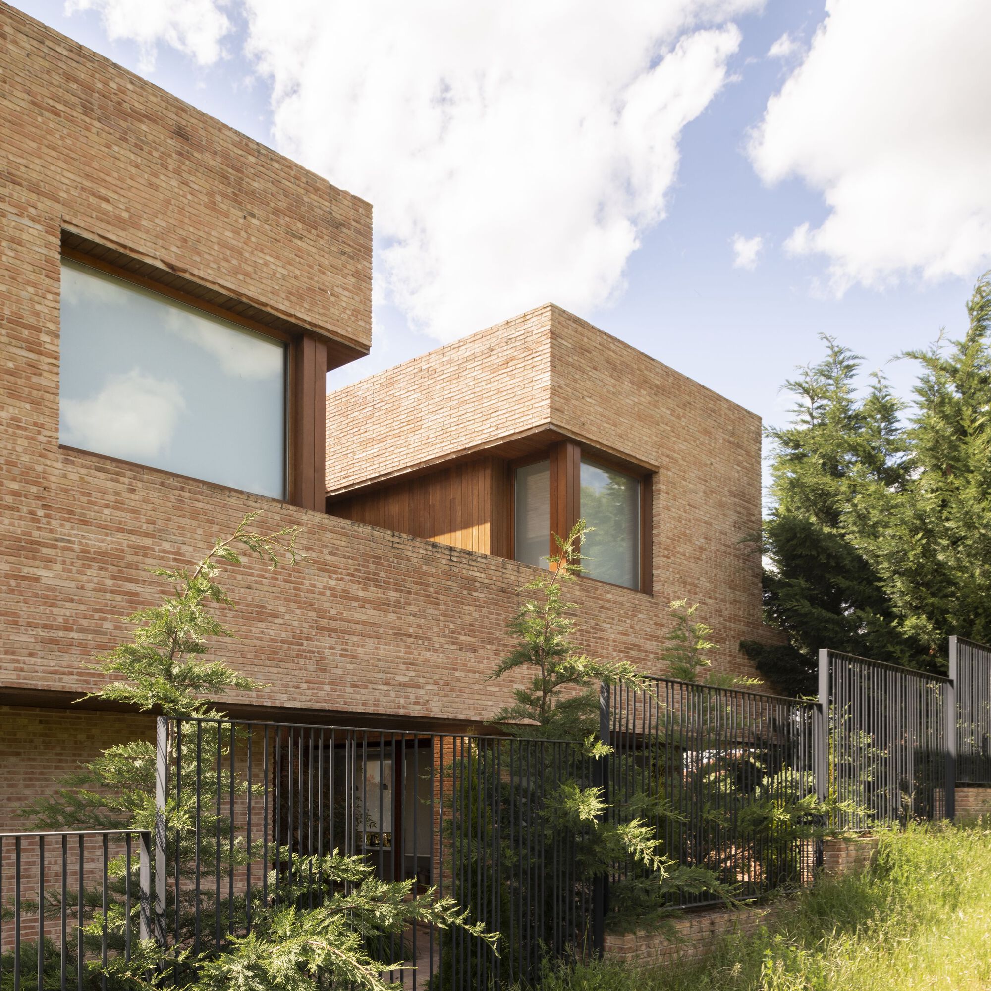 Stacked brick volumes with timber window recesses above a metal railing and young trees