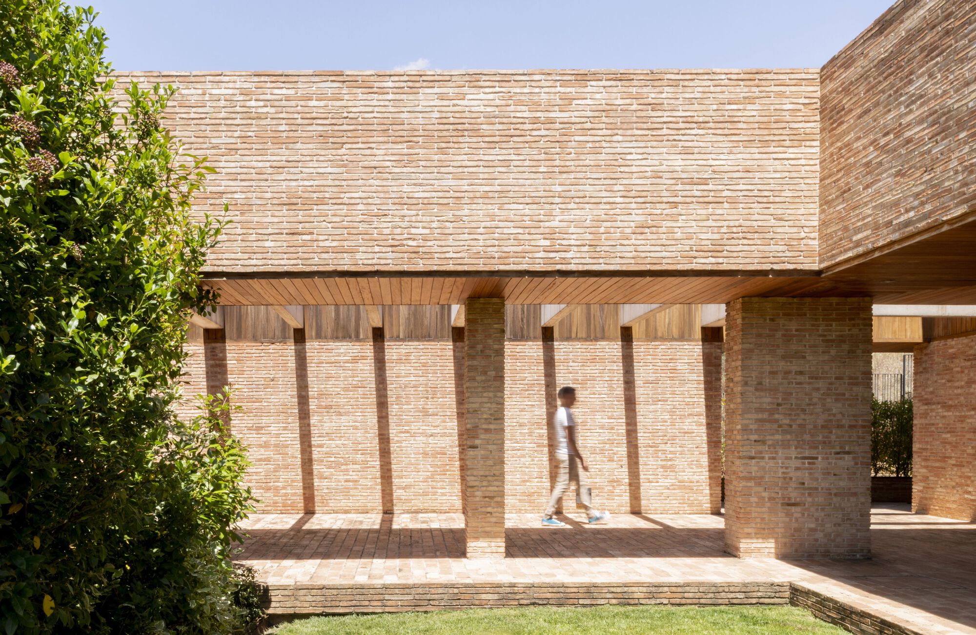 Covered walkway with timber ceiling and brick columns as a figure walks through midday light