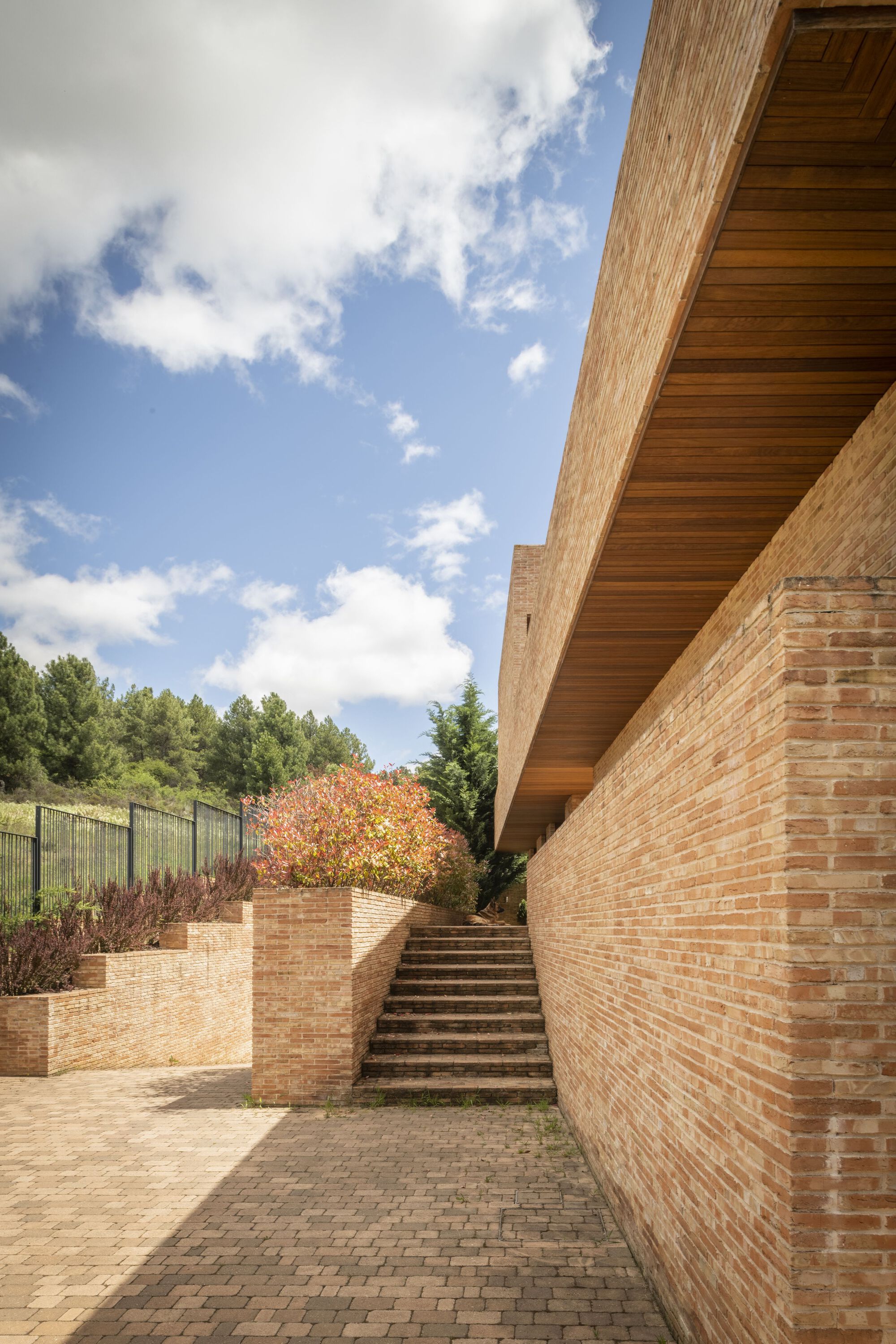 Brick-paved courtyard with stepped planters and timber-lined roof soffit beneath a blue sky with clouds