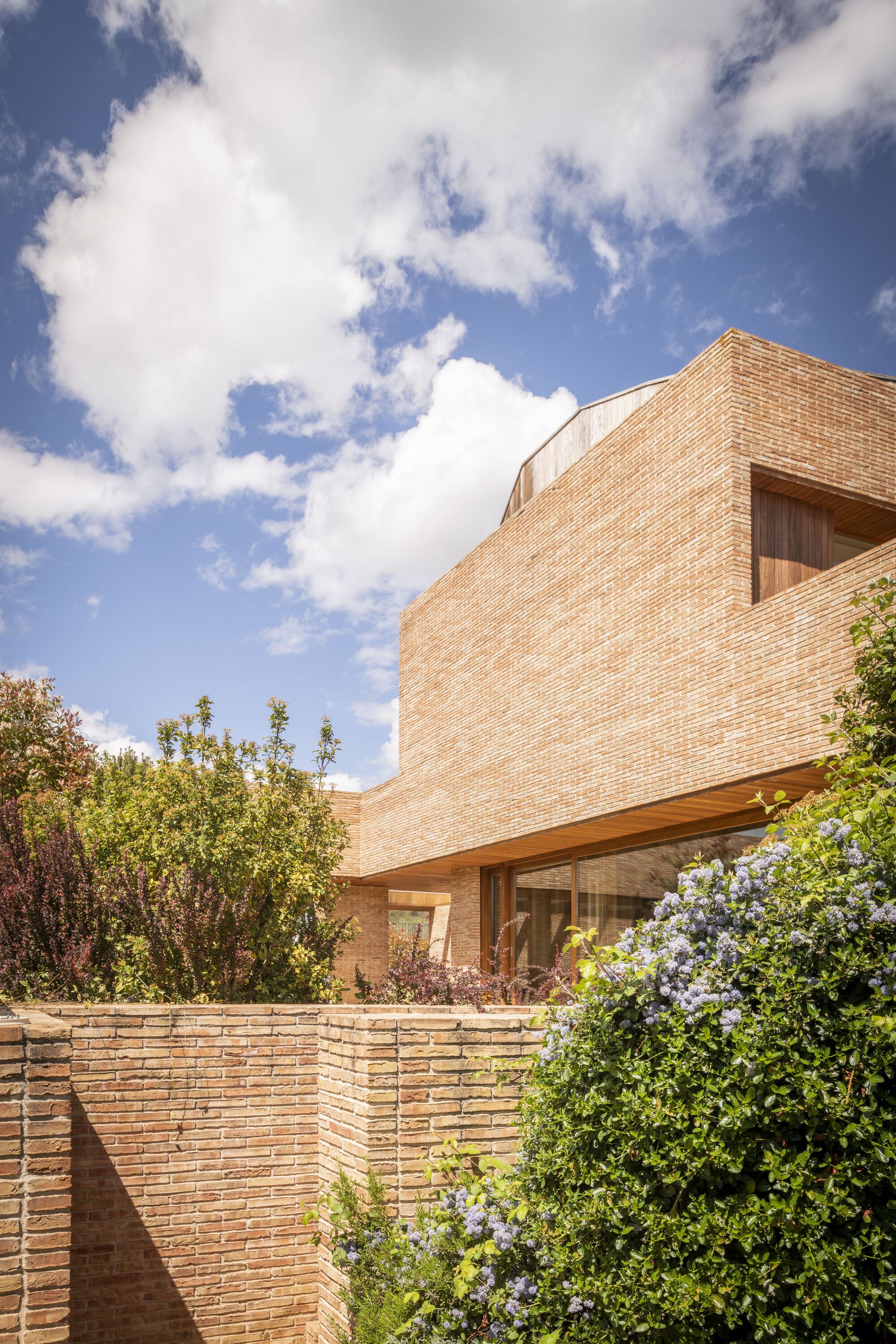 Two-story brick facade with timber soffit and glazed opening above a low retaining wall with flowering shrubs