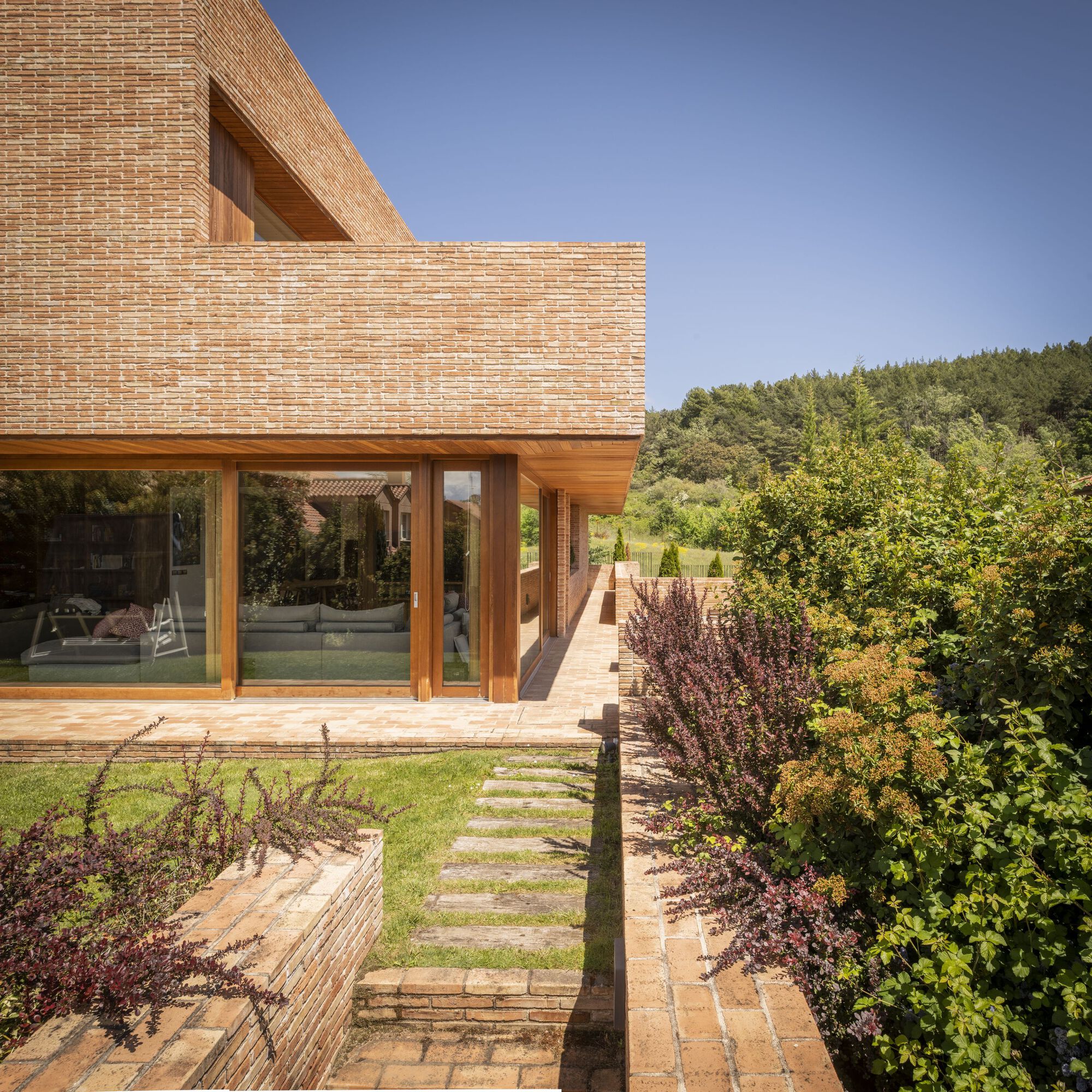 Garden view showing timber-framed glazing beneath brick volumes and stepping stones through planted beds