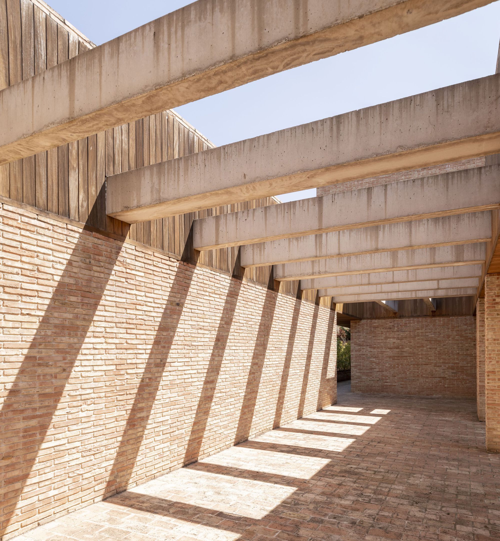 Covered brick walkway with exposed concrete beams casting diagonal shadows across the paved floor