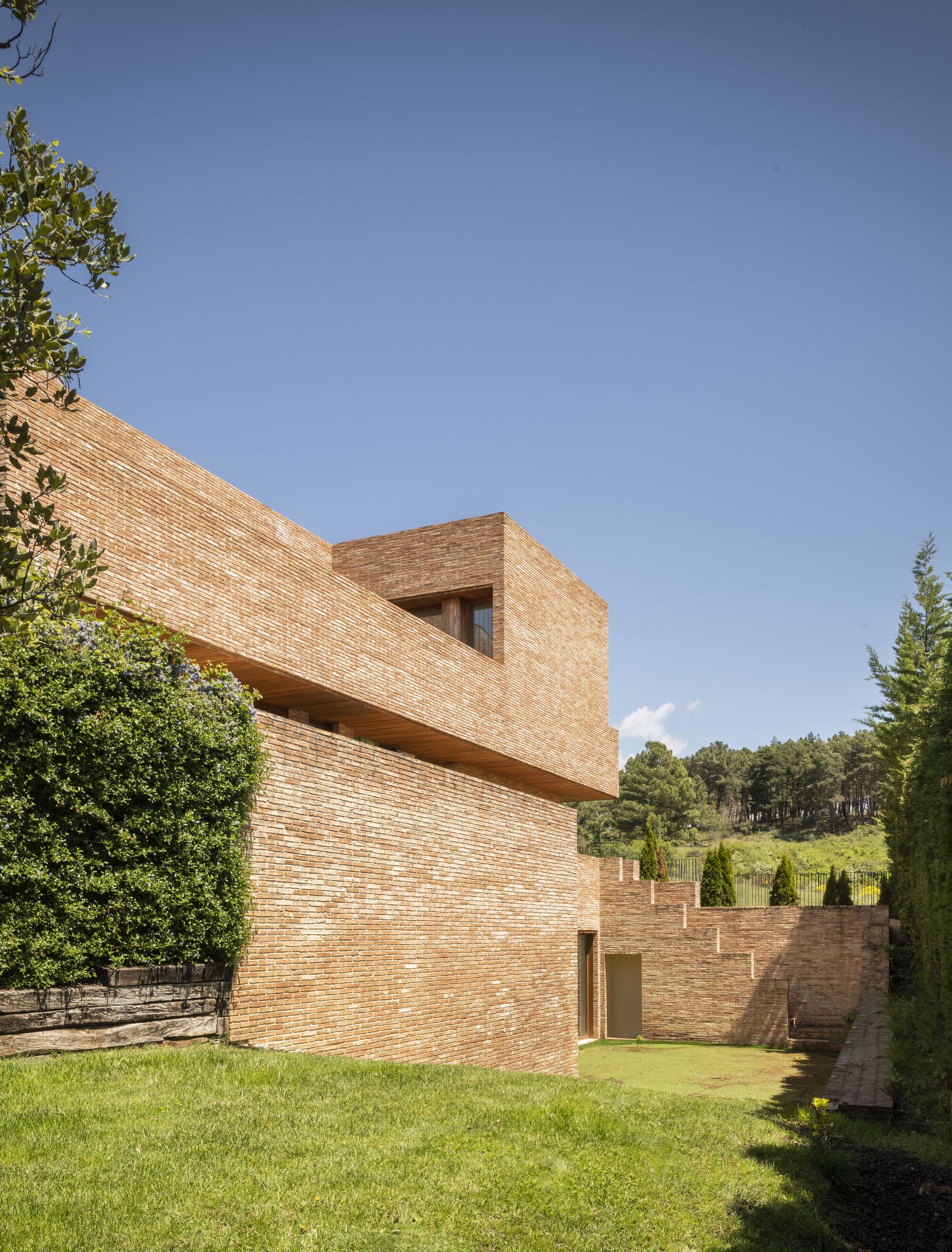 Stacked brick volumes with a terrace opening framed by a lawn and surrounding trees