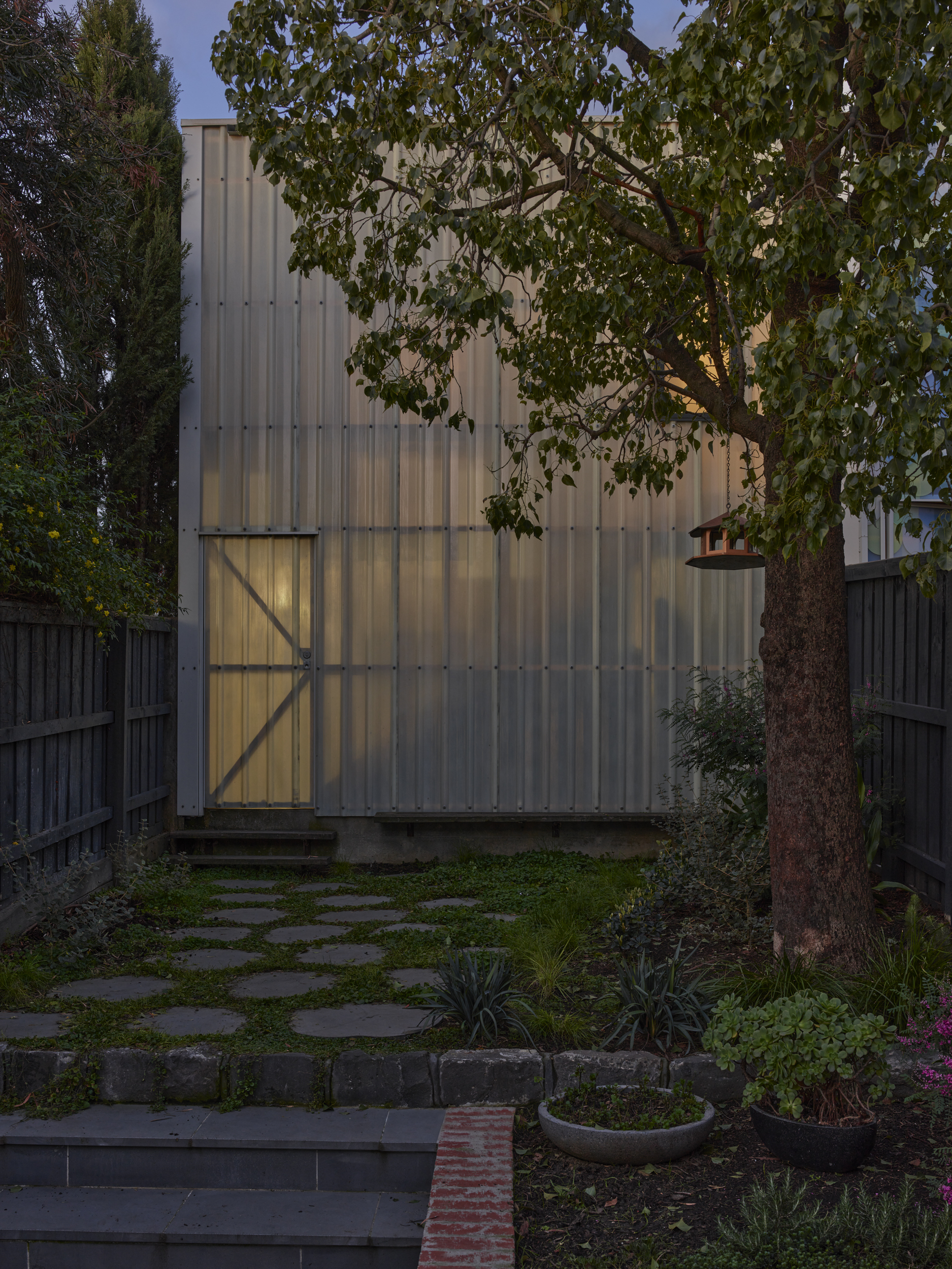 Corrugated metal facade with illuminated doorway framed by overhanging tree branches at dusk