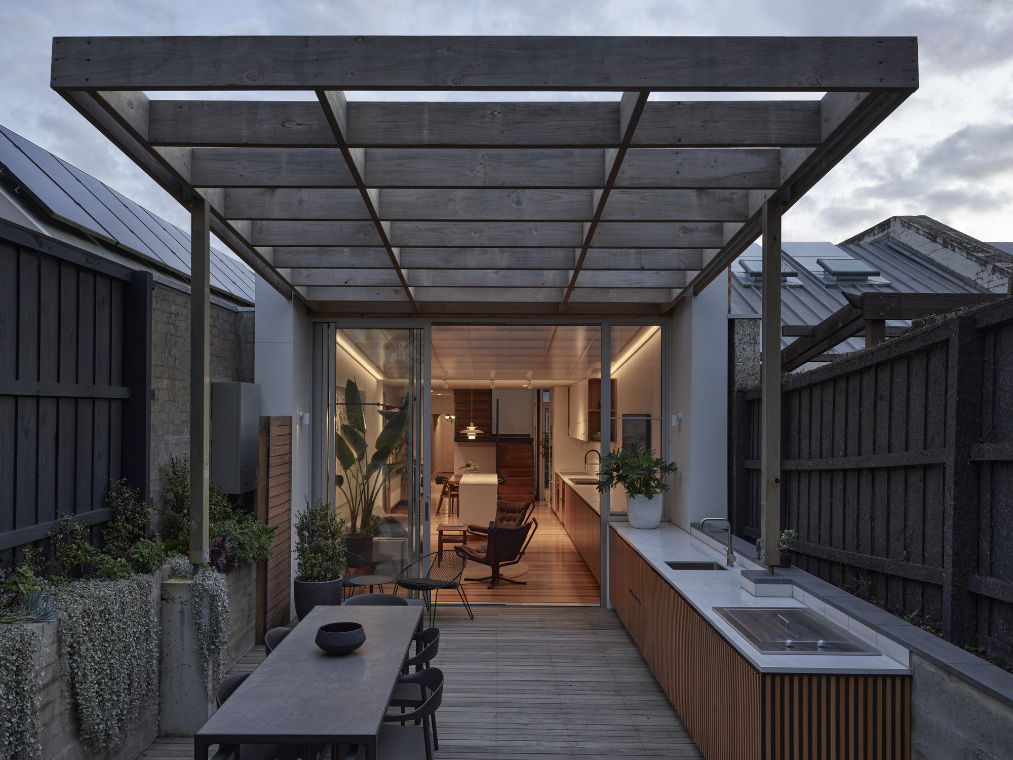 Rear courtyard with pergola, outdoor sink, and decking leading to an illuminated interior at dusk