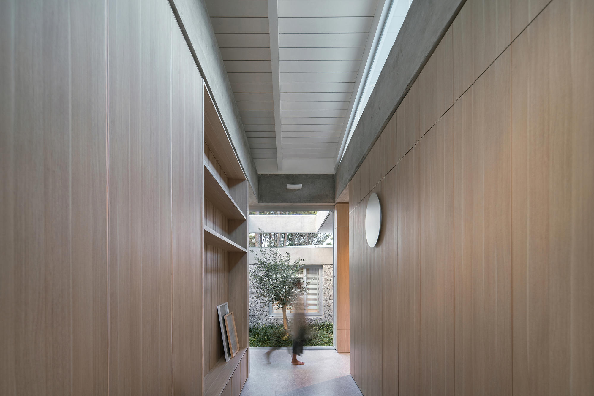 Hallway with timber-lined walls and ceiling leading to a planted courtyard as a figure passes through