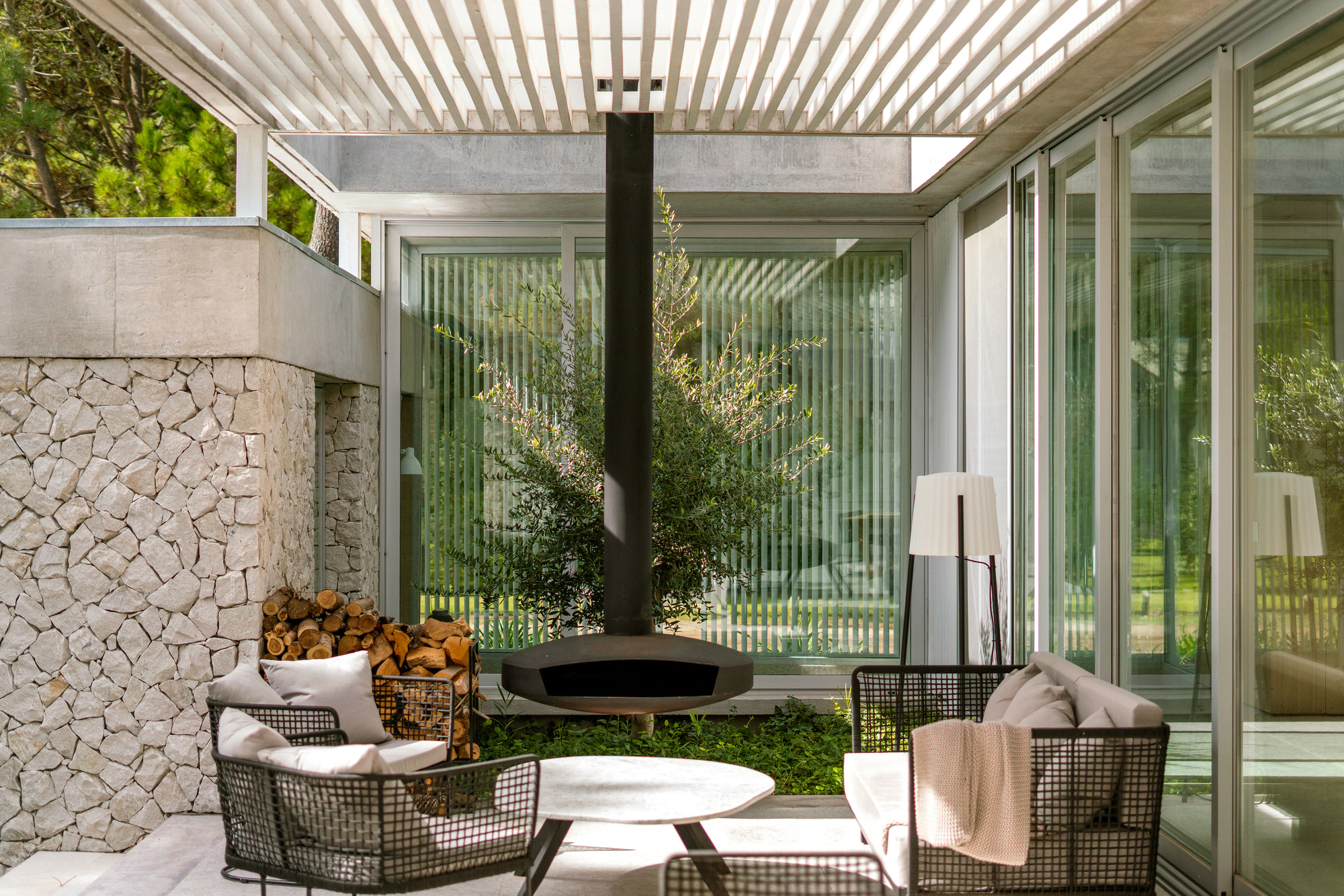 Covered terrace with slatted pergola, suspended fireplace, and stone wall beside planted olive tree