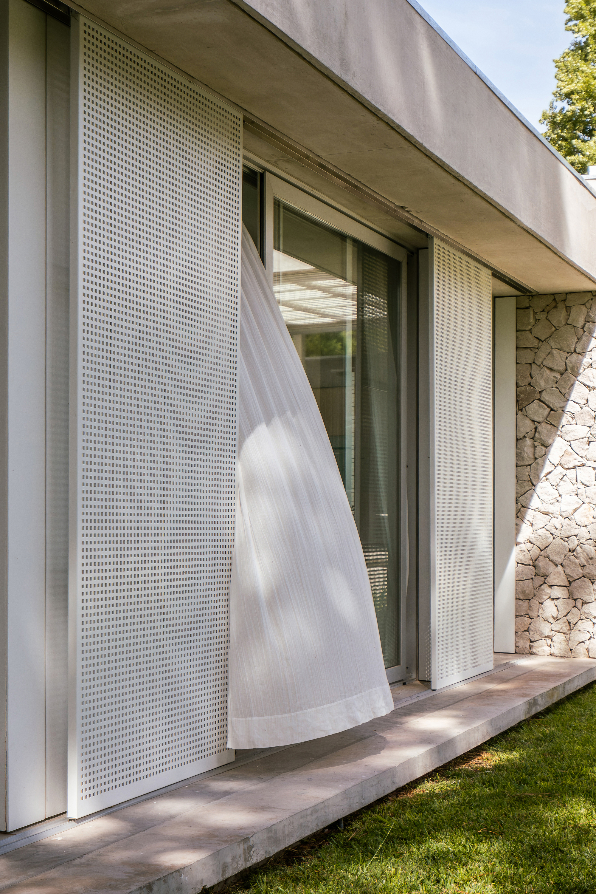 Perforated metal screen panel lifted by breeze beside glass sliding door and stone wall corner