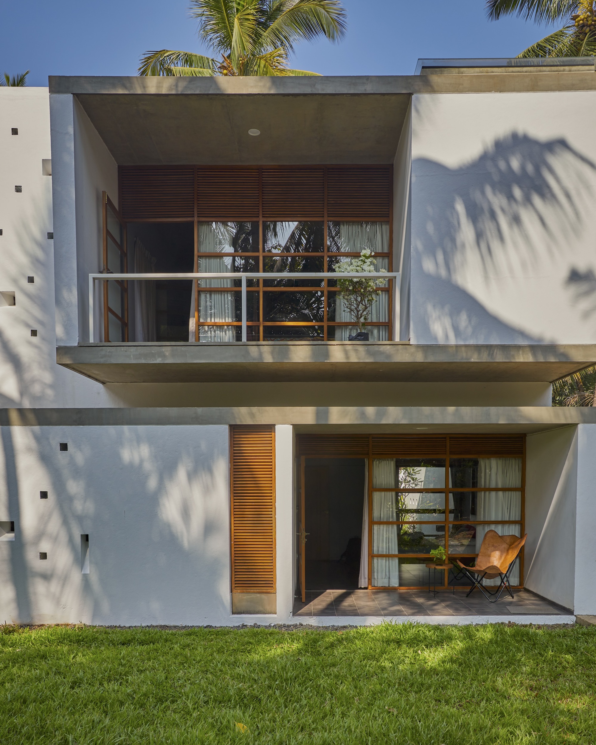 Two-story facade with cantilevered balconies and timber louvered panels framed by palm shadows