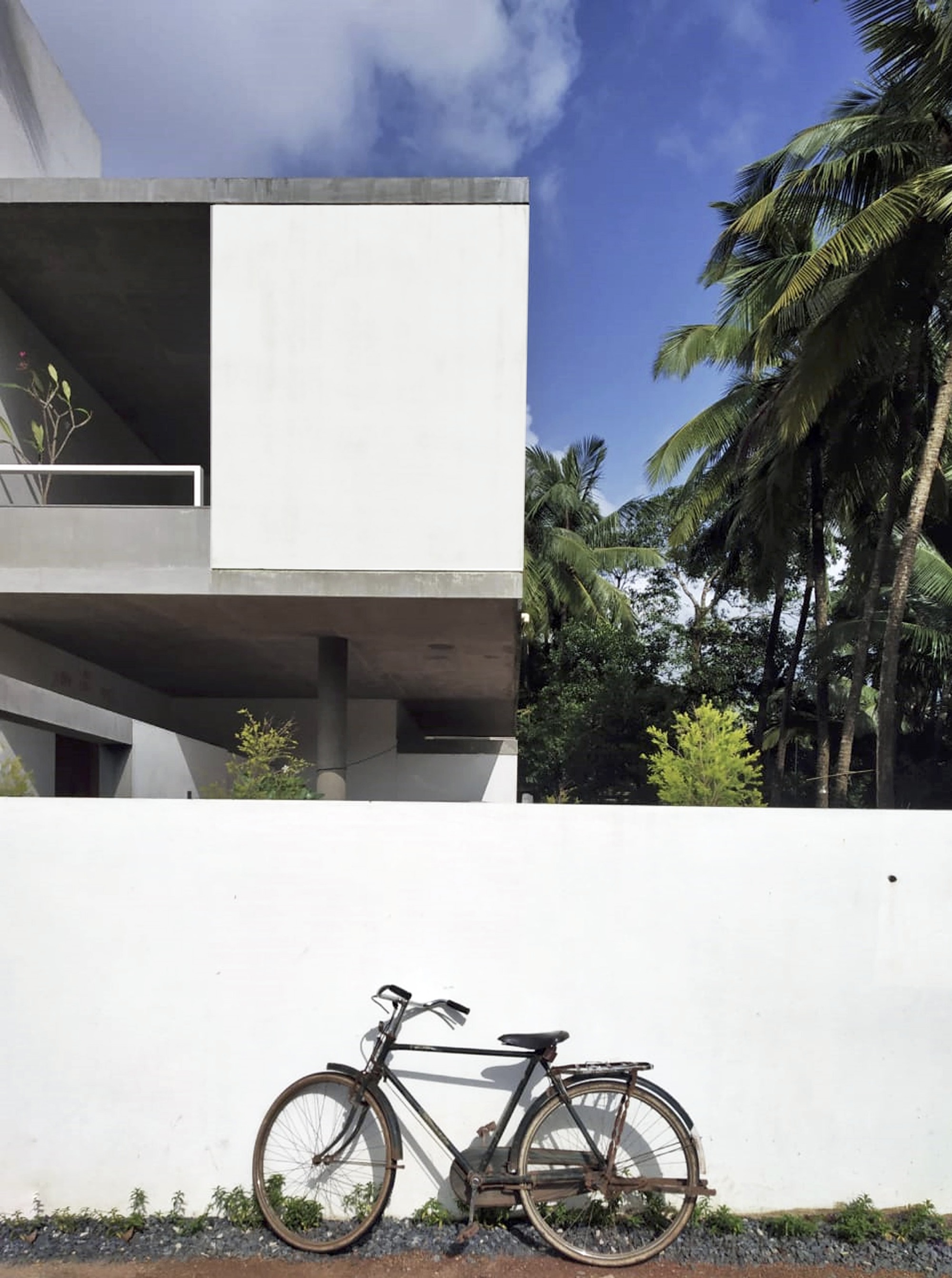 White stucco boundary wall with parked bicycle below cantilevered concrete volumes and palm trees