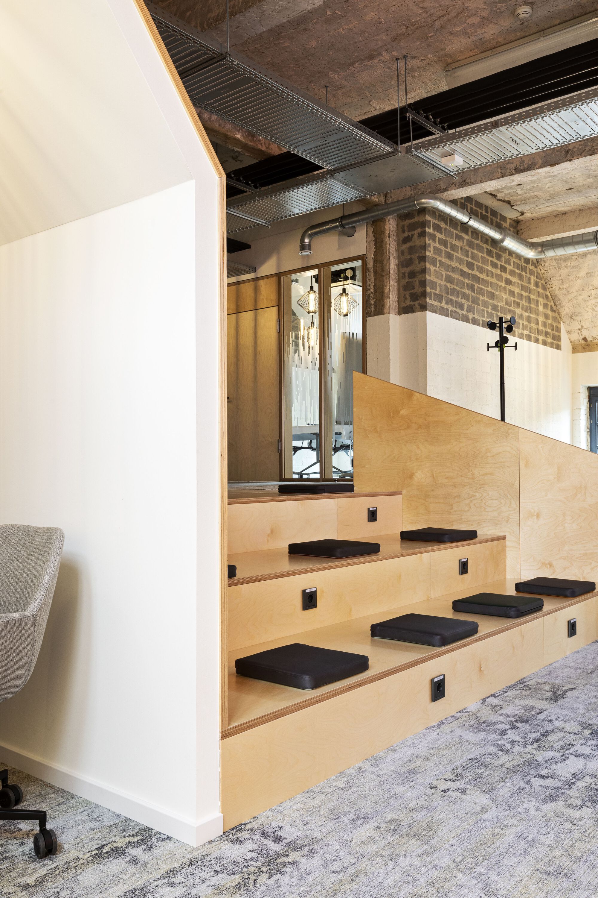Plywood staircase with black cushioned seating treads beneath exposed concrete ceiling and mechanical ducts