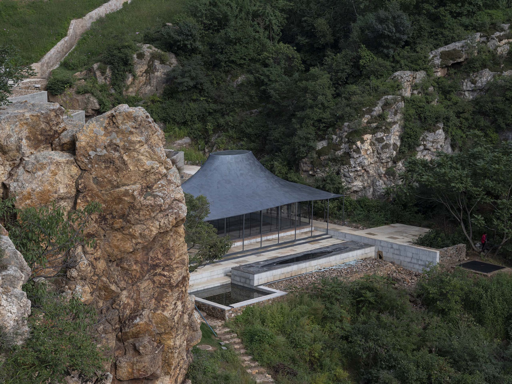 Aerial view of the glazed pavilion with curved metal roof nestled between limestone cliffs and vegetation