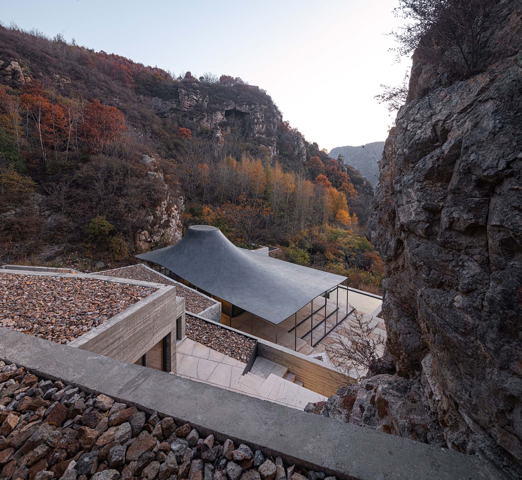 Pavilion with sweeping metal roof set within a narrow gorge framed by autumn foliage at dusk