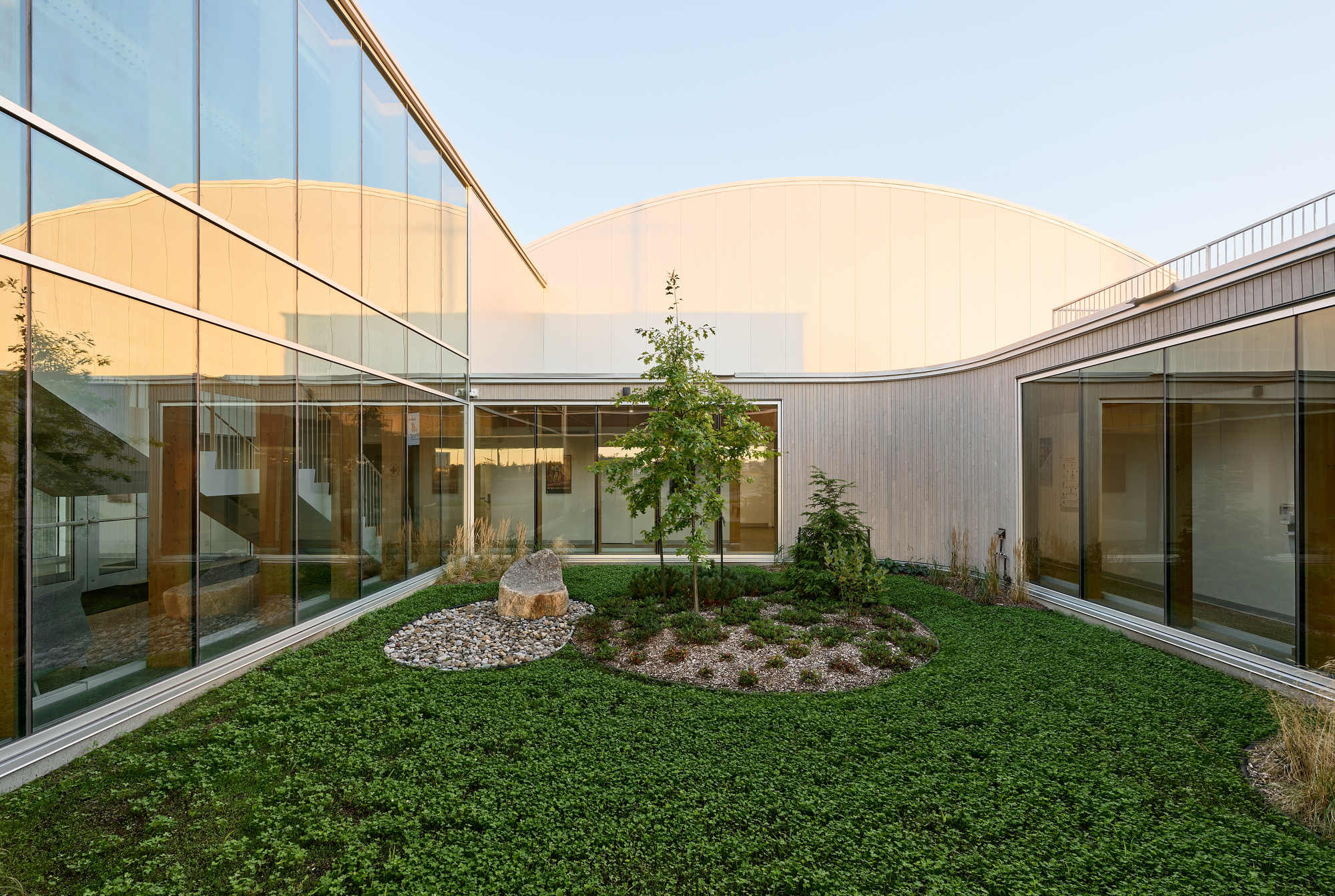 Planted courtyard with circular groundcover bed and trees surrounded by glass-walled corridors under an overcast sky