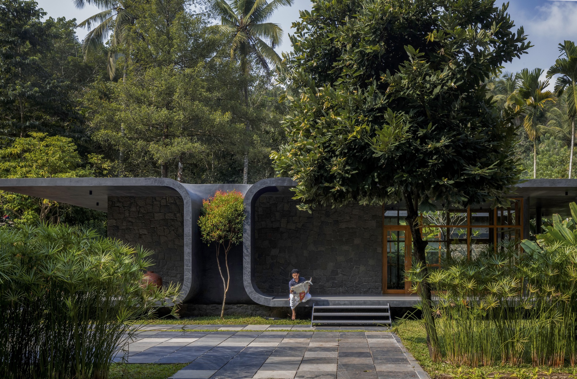 Entry courtyard with curved concrete facade, planted bed with young tree, and a figure seated on steps