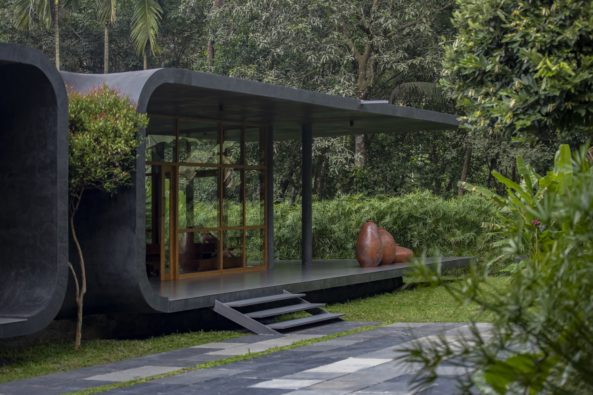 Glass pavilion with timber framing and curved concrete roof edge set in a lawn with tropical vegetation