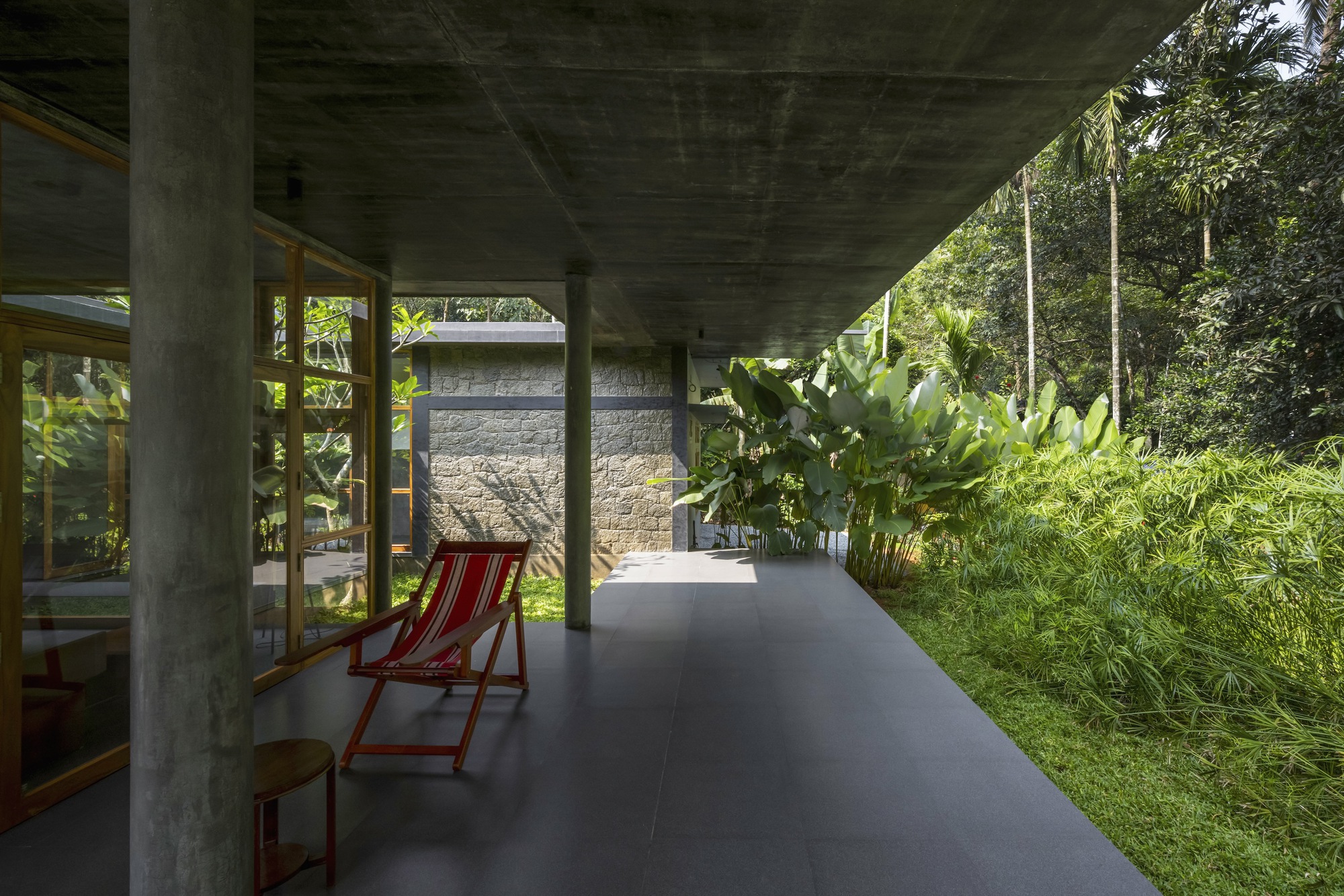 Covered passage with concrete columns and a red deck chair facing a stone wall and palms