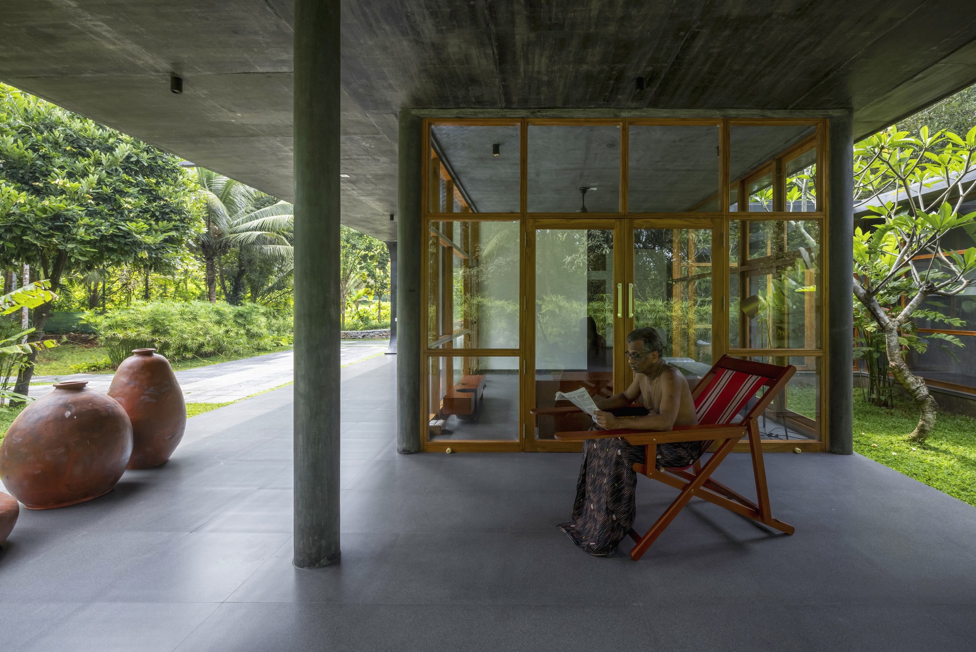 Covered terrace with concrete columns and timber-framed glazing opening to tropical garden with person reading
