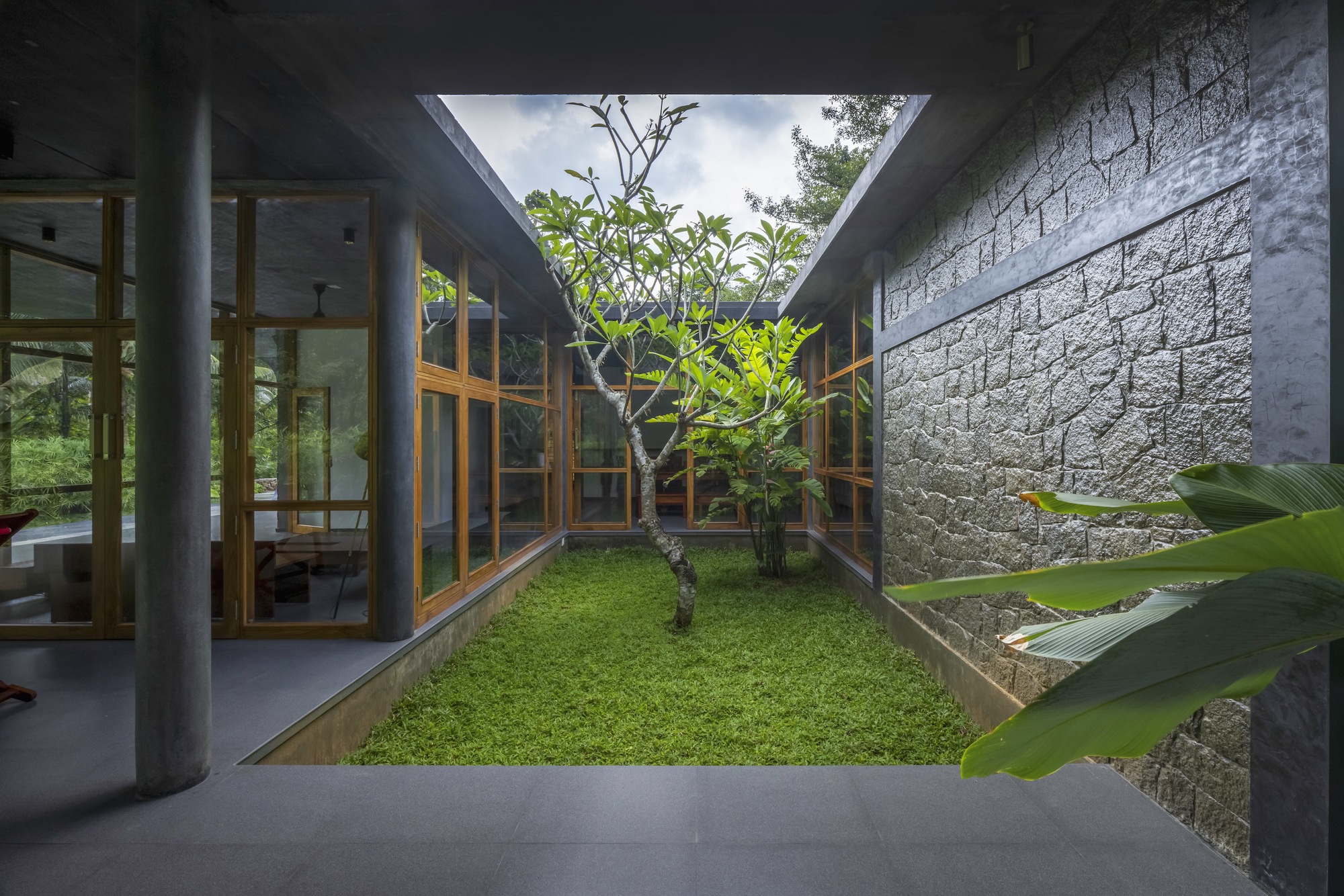 Courtyard with grass lawn and single tree framed by textured stone walls and timber window walls