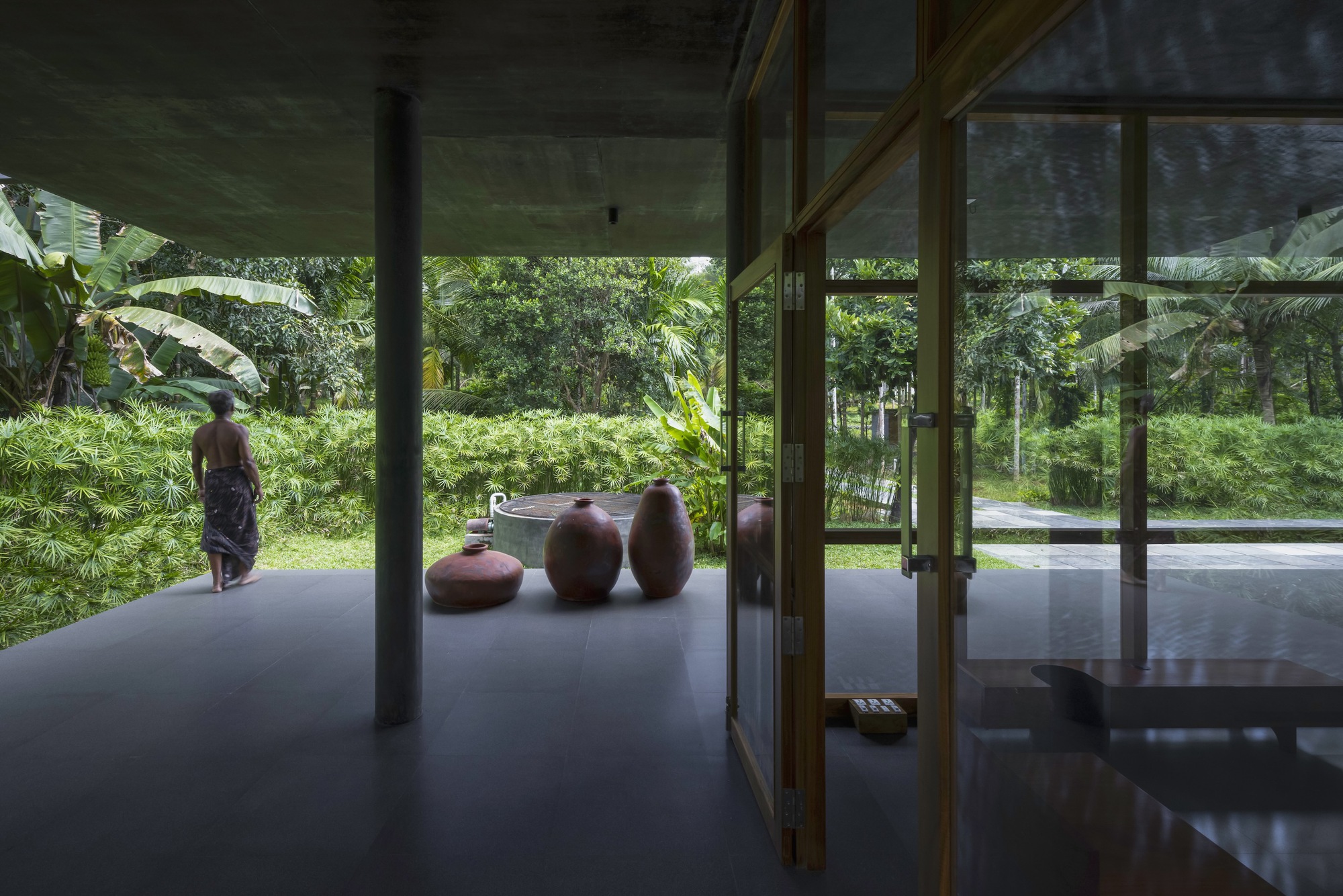 Open pavilion with polished floor and ceramic vessels looking toward garden with person standing outside