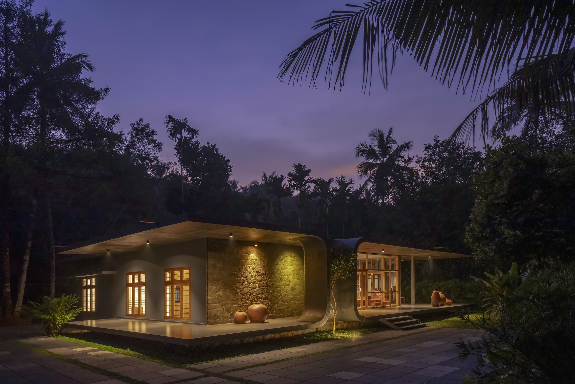 Curved roofline with illuminated stone wall and glazed entrance at dusk beneath palm trees