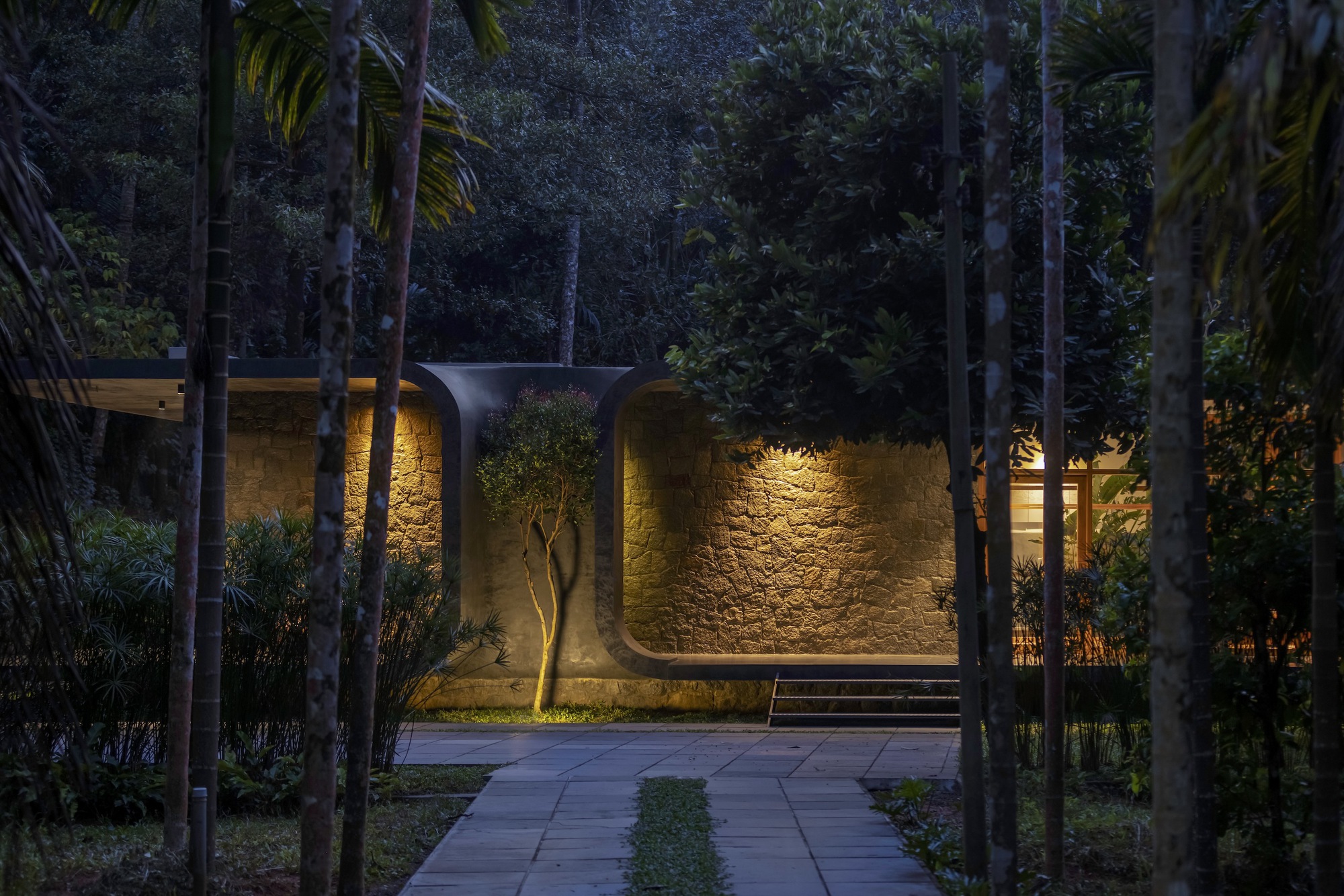Night view of the illuminated stone facade with arched niches framed by palms and planted approach