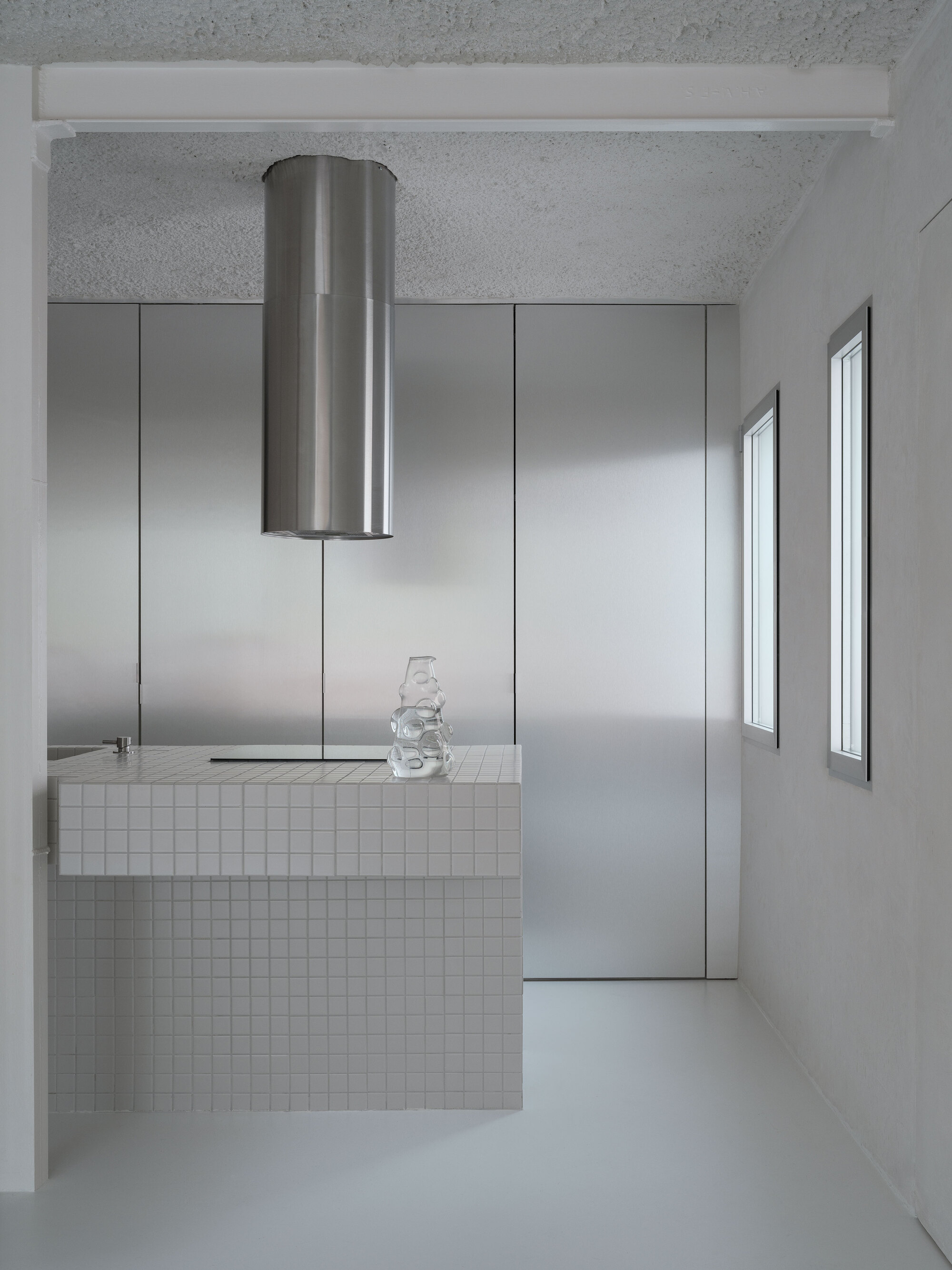 Kitchen island clad in white mosaic tile with cylindrical steel extractor above and glass vessel