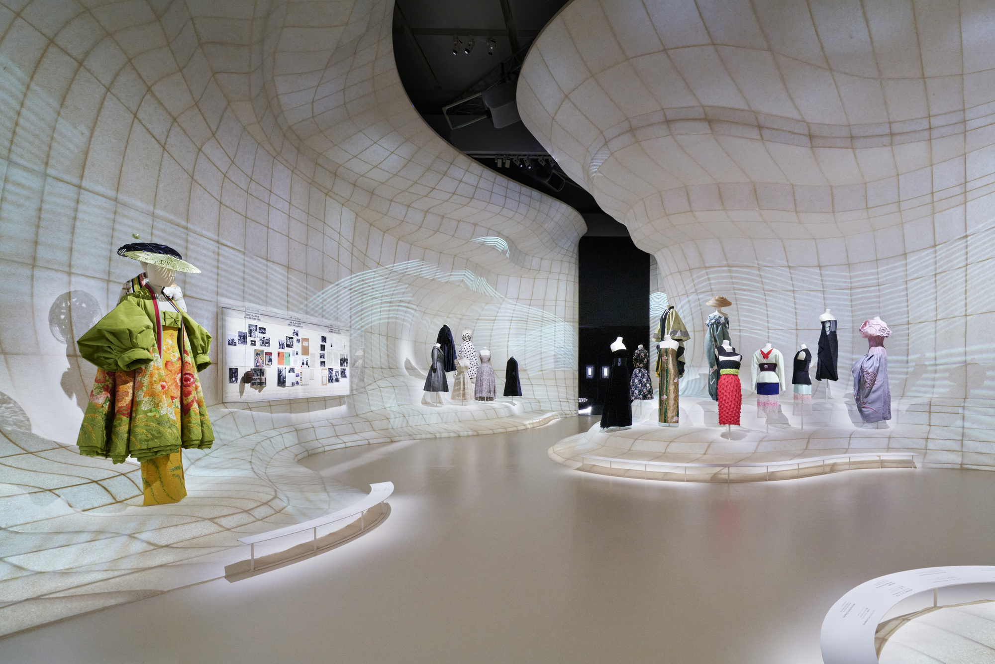 Curved white grid-patterned walls enclosing a gallery space with fashion mannequins on display platforms