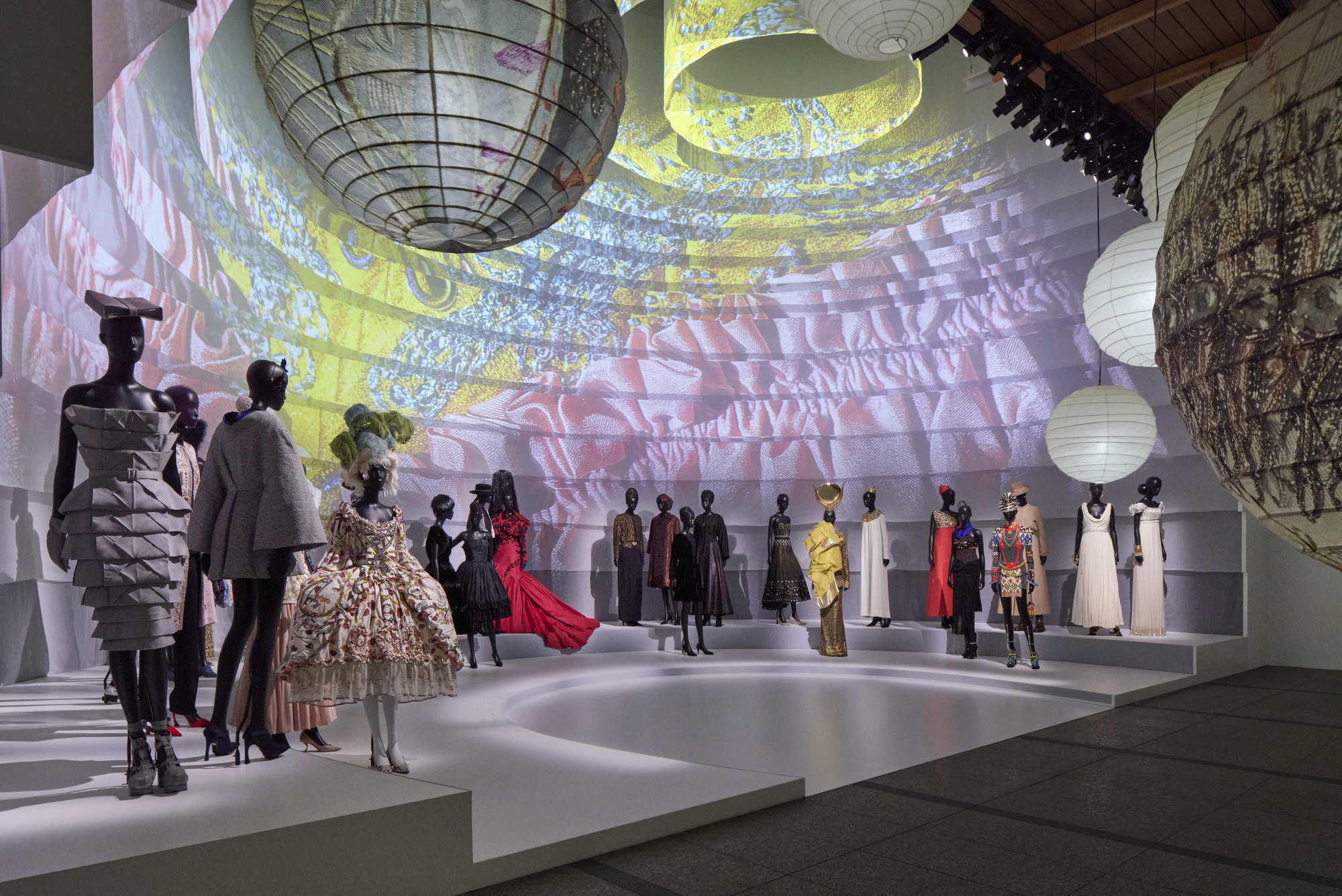 Gallery space with suspended lanterns and projected imagery across white stepped platforms holding fashion displays