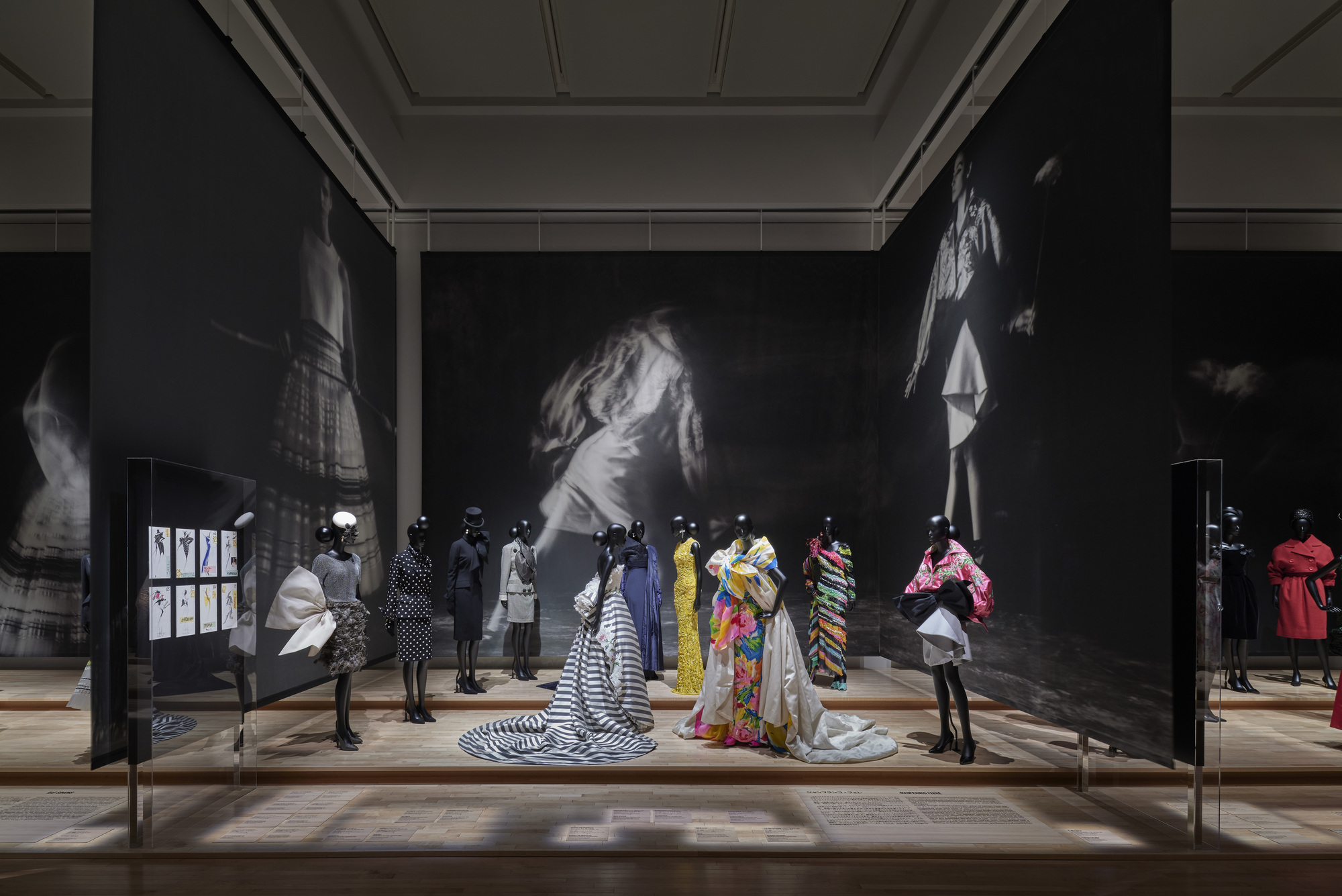 Gallery room with illuminated mannequins in colorful garments beneath large-scale black-and-white portrait projections