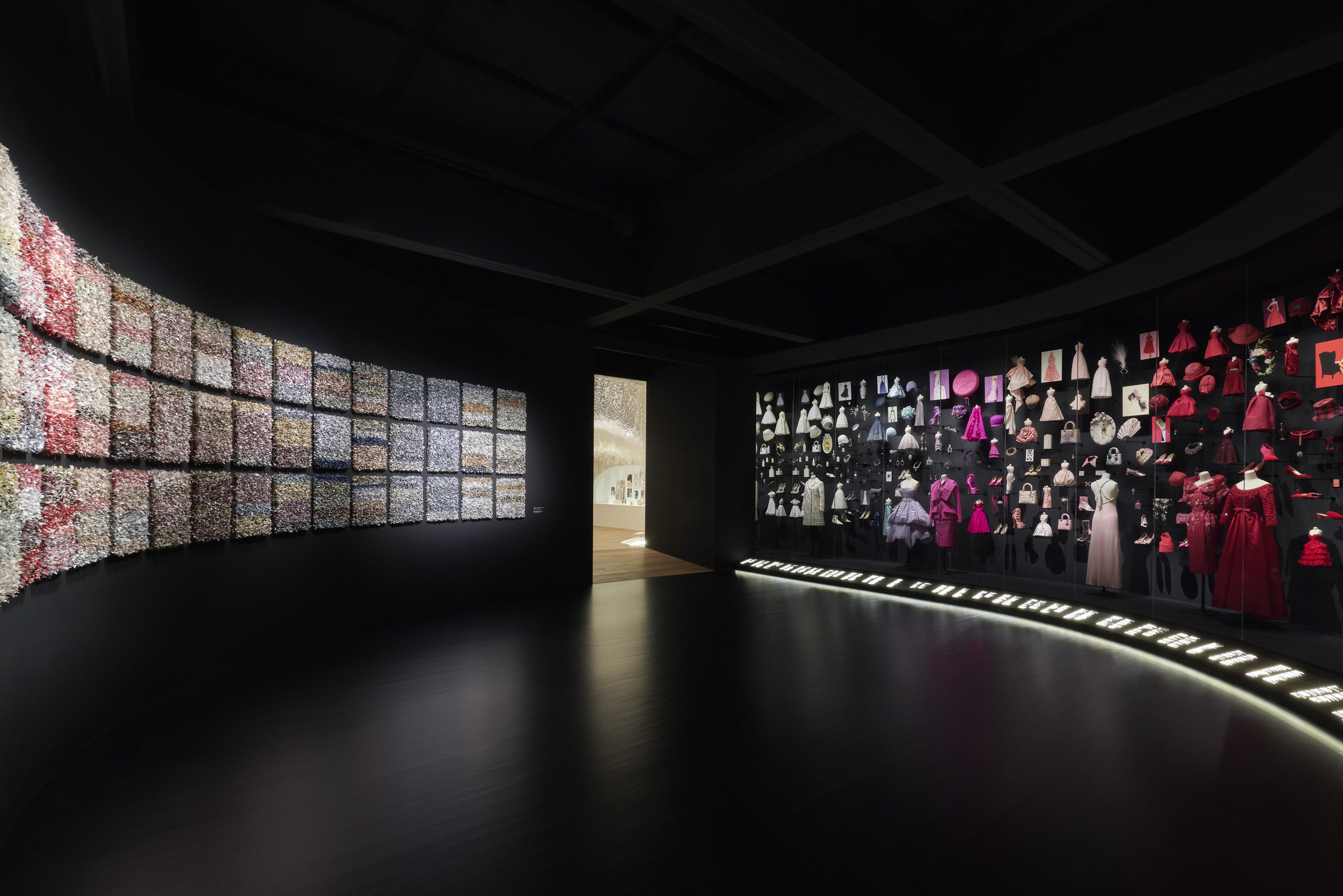 Curving exhibition space with backlit image grid and wall of garments under a black coffered ceiling