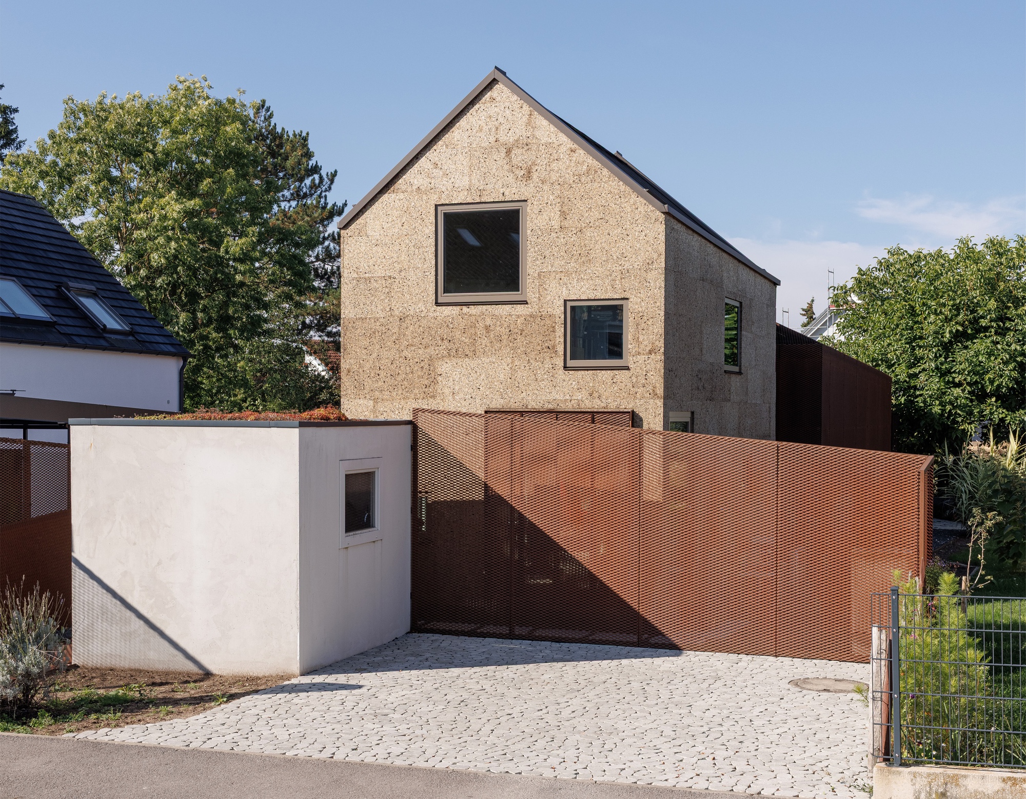 Street view of the cork-clad gable facade with perforated copper gate and white stucco entry volume
