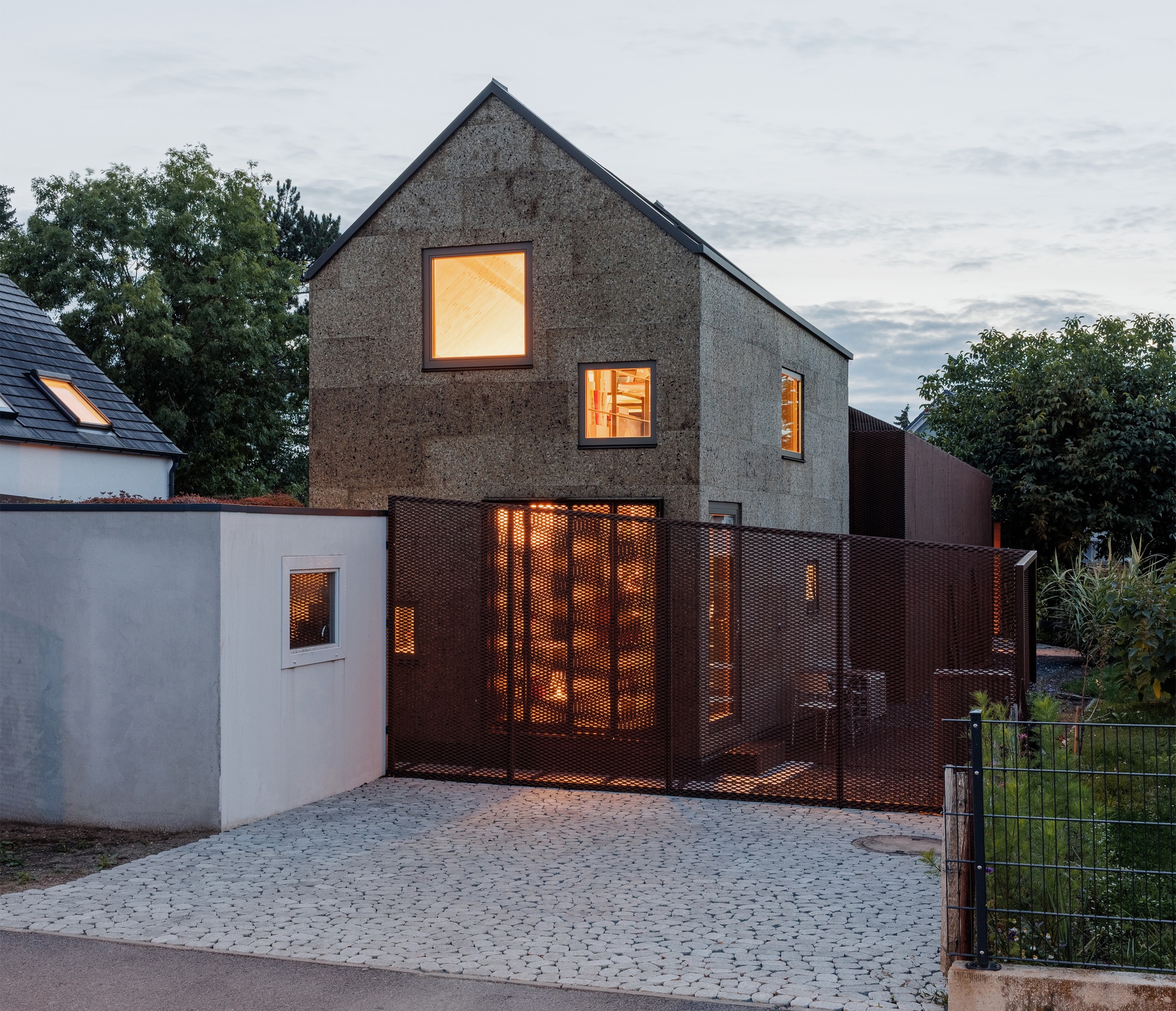 Street facade at dusk with glowing windows and illuminated interior visible through perforated copper gate