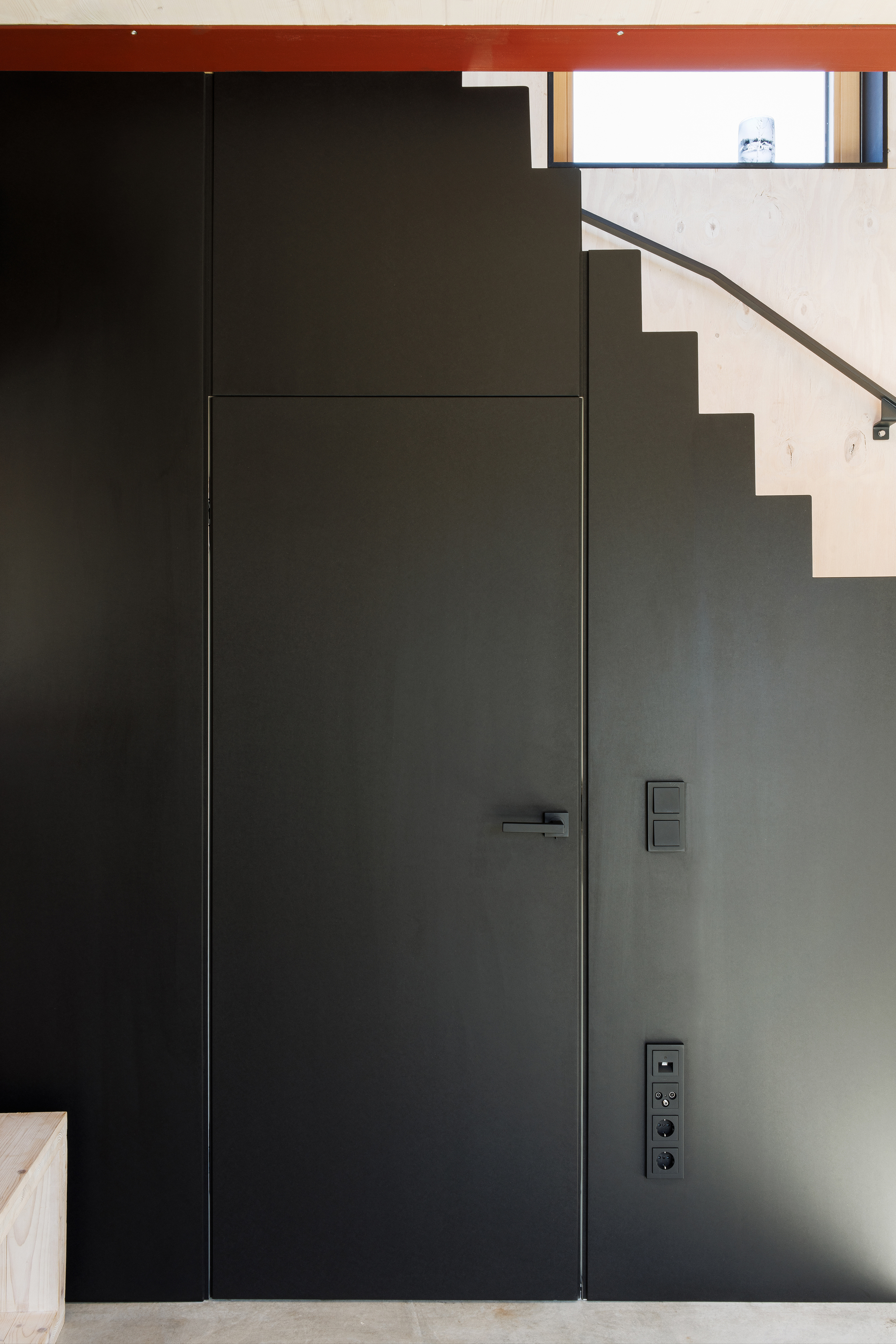 Flush black metal door set into grey wall beneath visible stair treads and red ceiling beam