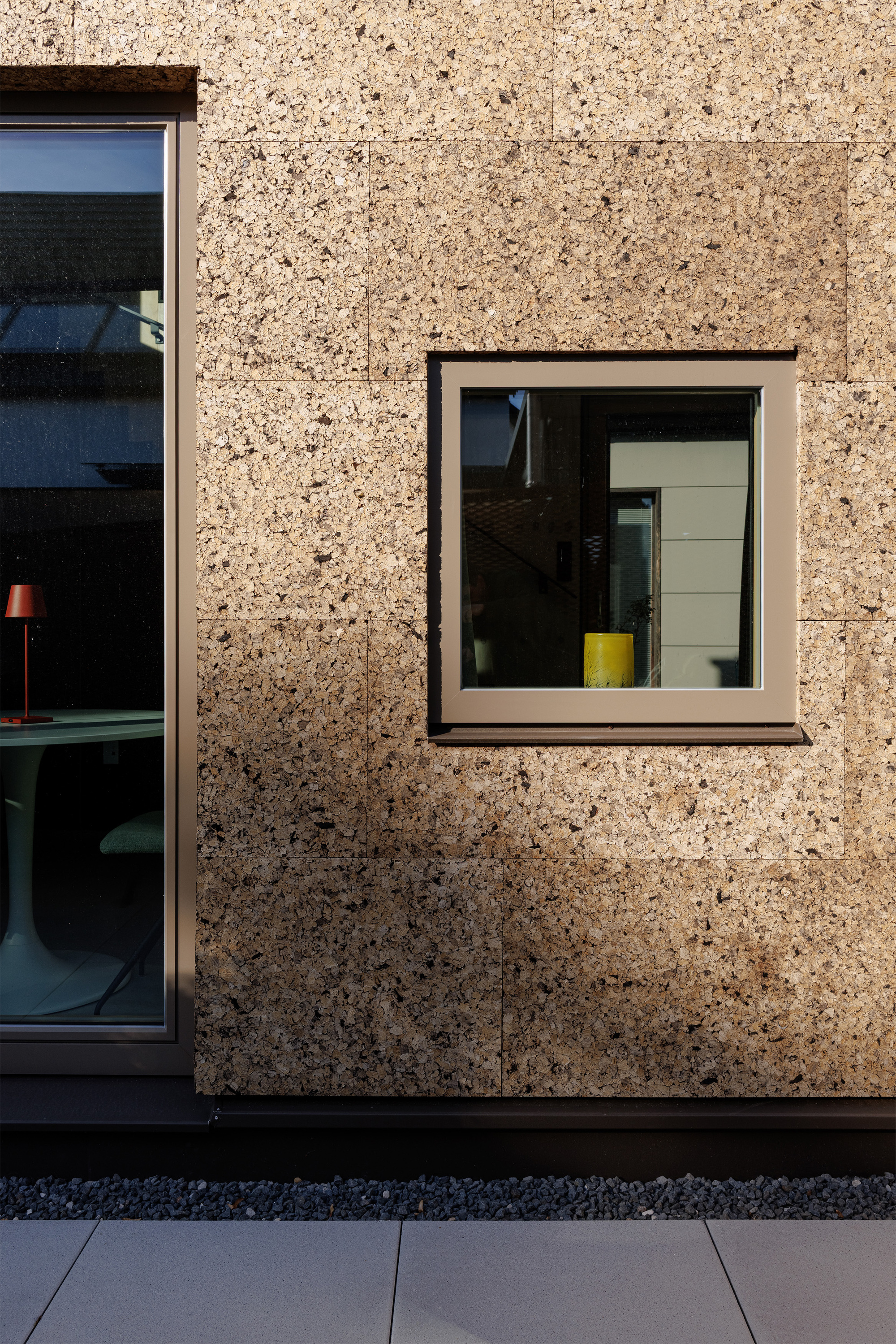 Cork aggregate facade with two aluminum-framed windows revealing interior spaces and a yellow stool