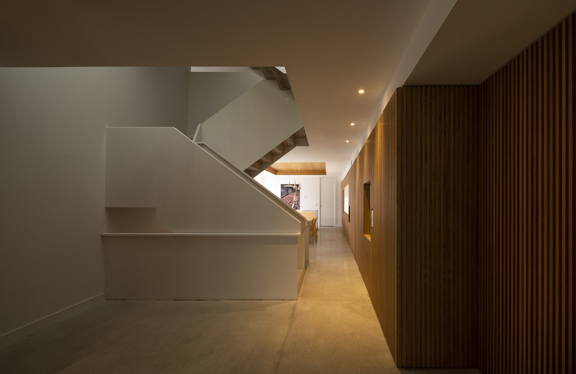 Ground floor hallway with vertical timber slat wall and white plaster staircase under recessed lighting
