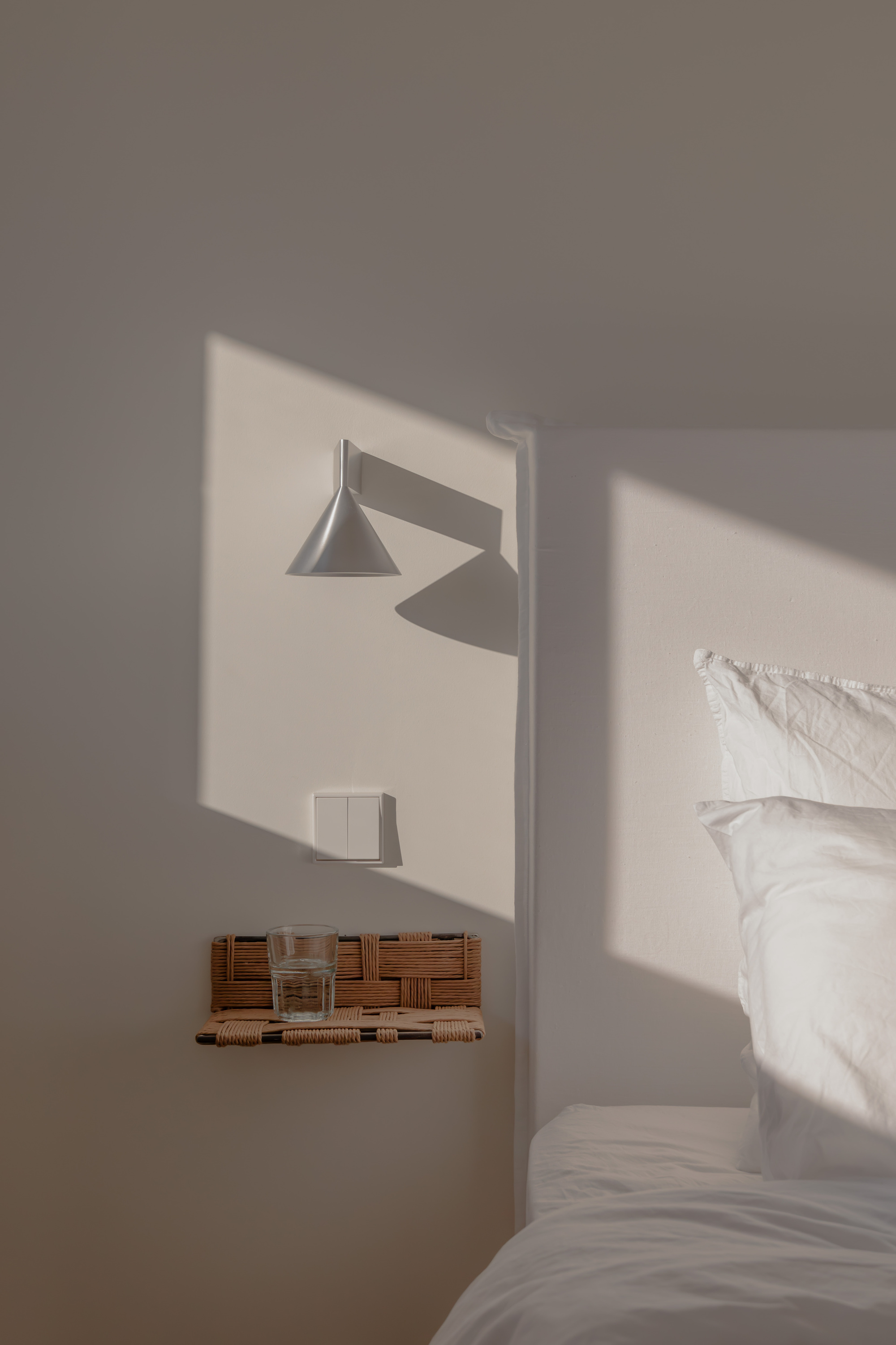 Bedroom niche with wall-mounted lamp and woven shelf casting afternoon sunlight across white surfaces