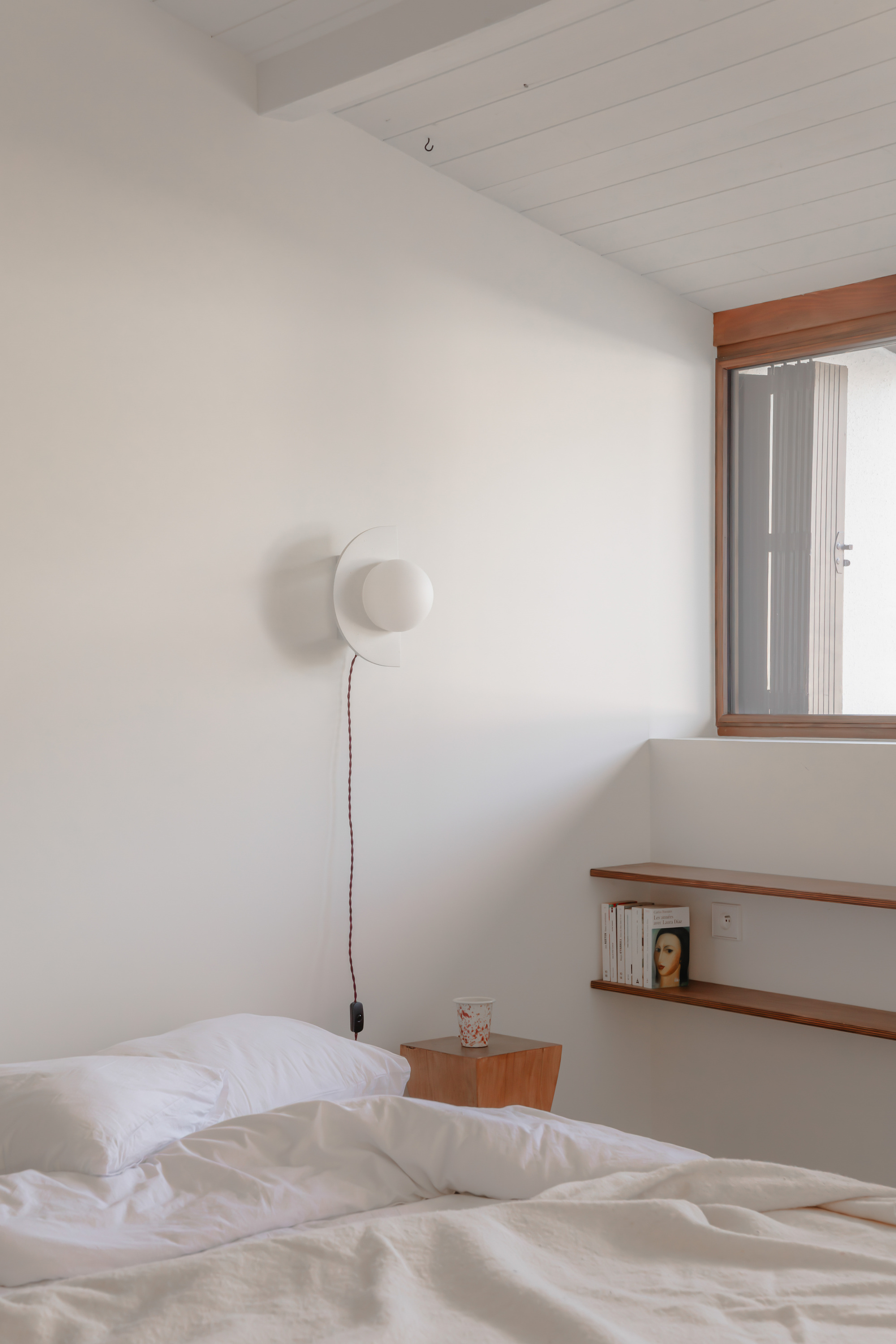 Bedroom corner with timber window frame, wall sconce, open shelving and white painted tongue-and-groove ceiling