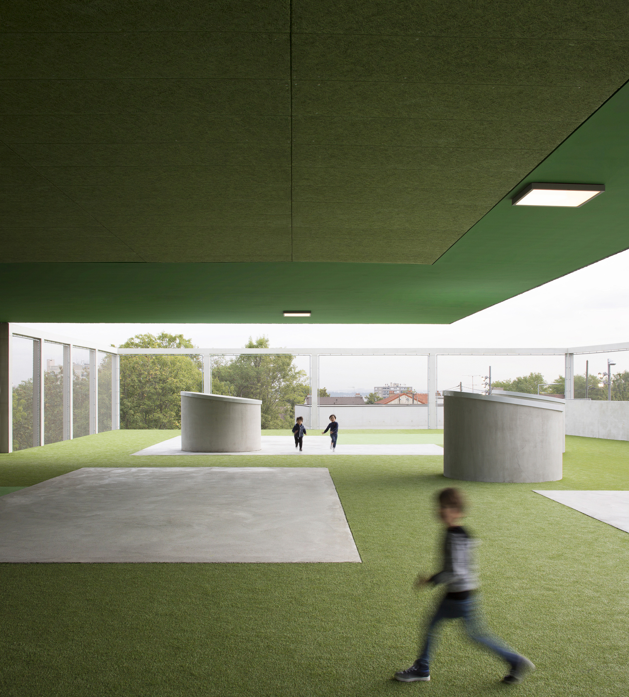 Covered play area with green soffit, cylindrical concrete elements, and children on artificial turf