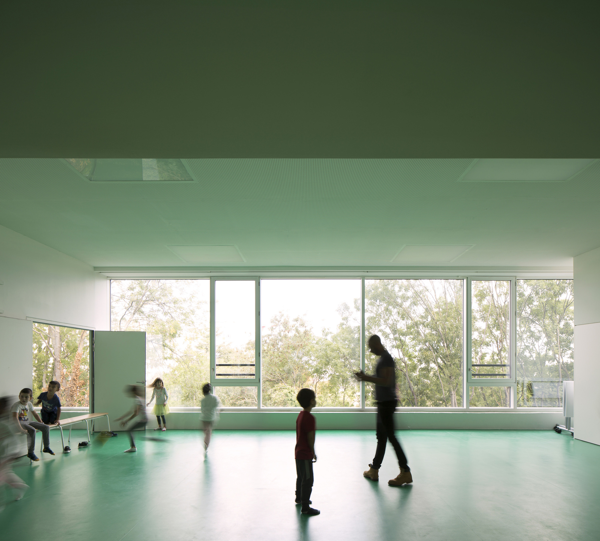 Interior classroom with green flooring and full-height windows as children move through diffused daylight