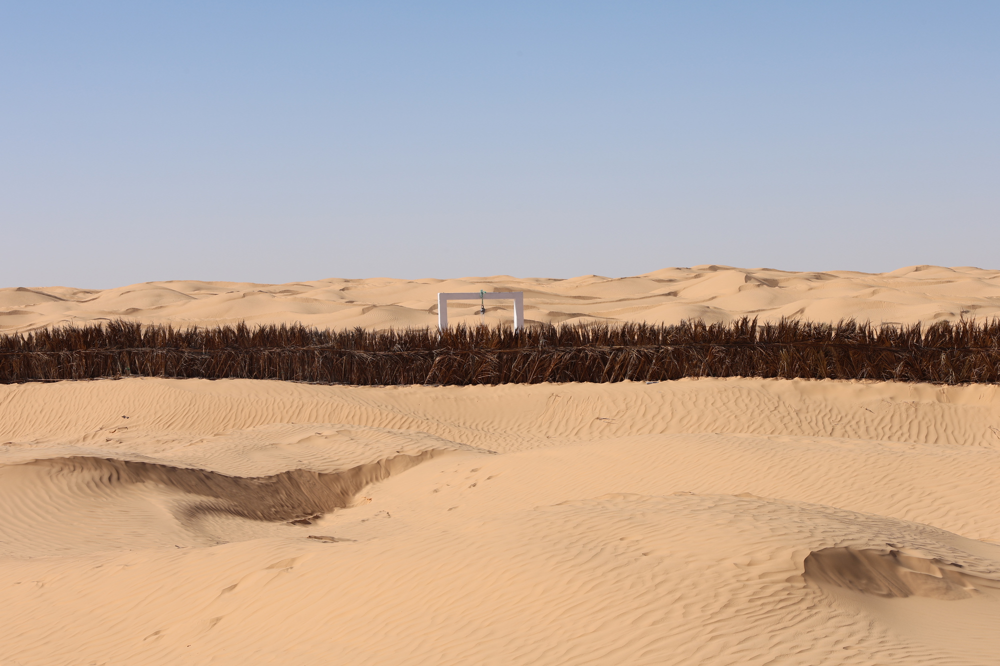 Distant view of a freestanding portal frame beyond a palm frond fence in rolling sand dunes