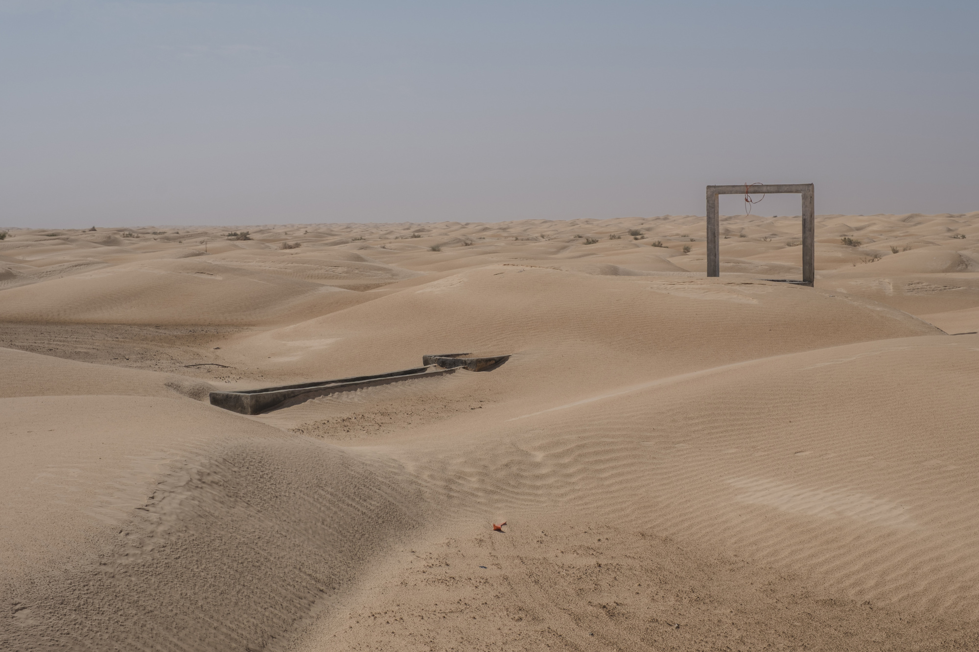 Concrete portal structure and partially buried water trough in windswept desert terrain under overcast sky