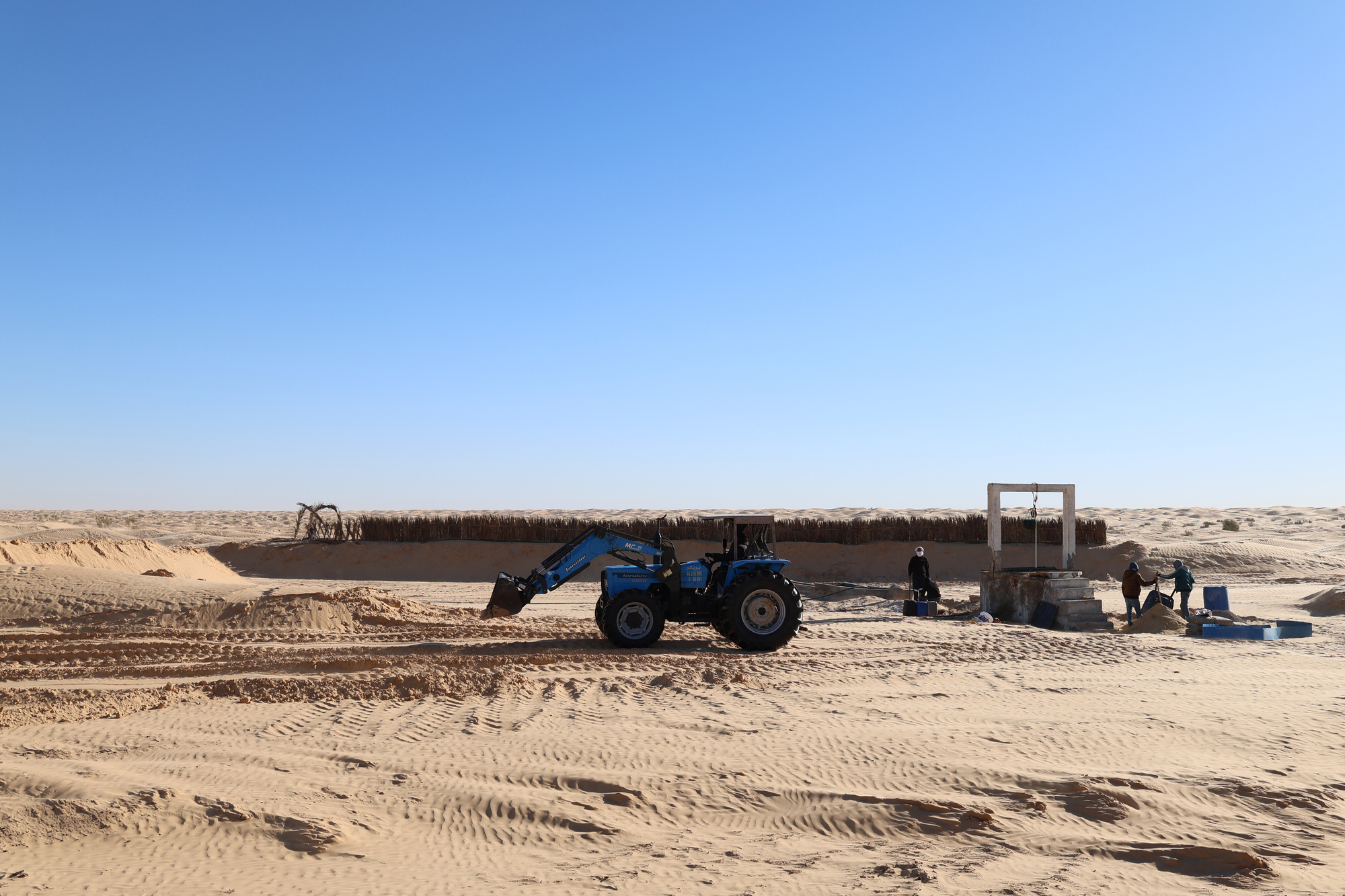 Construction view showing a blue tractor beside a palm frond wall and portal frame in sandy terrain