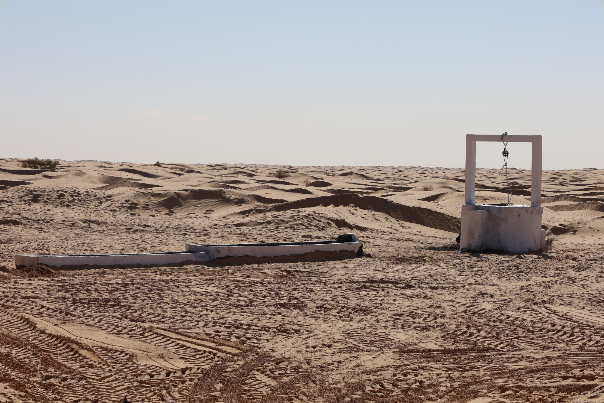 Concrete well structure with pulley system and adjacent water trough surrounded by tire tracks in sand