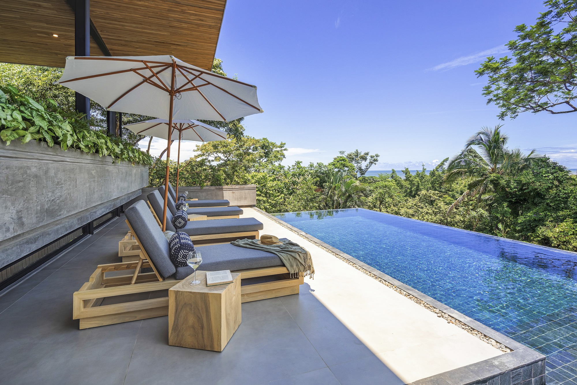 Infinity pool deck with timber loungers and white umbrella overlooking forested hills under blue sky