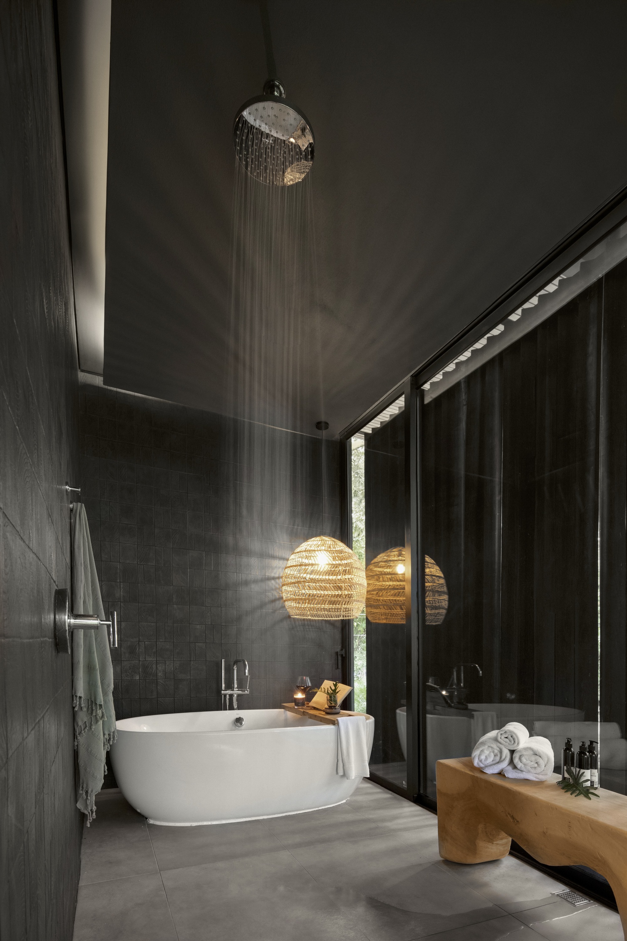 Dark tiled bathroom with freestanding tub under rainfall showerhead and woven pendant light