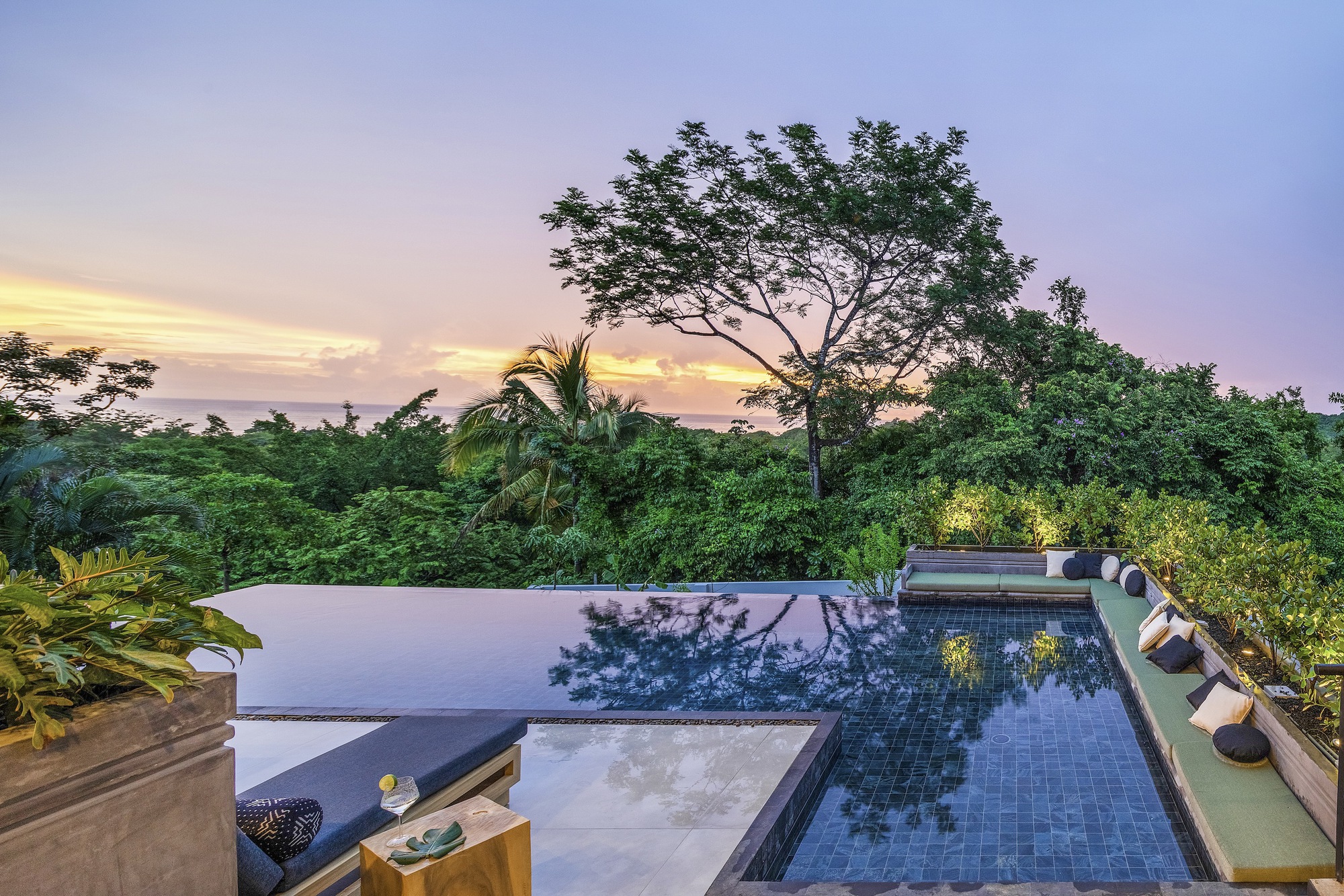 Infinity pool terrace with dark tile mosaic and lounge seating overlooking tropical canopy at dusk