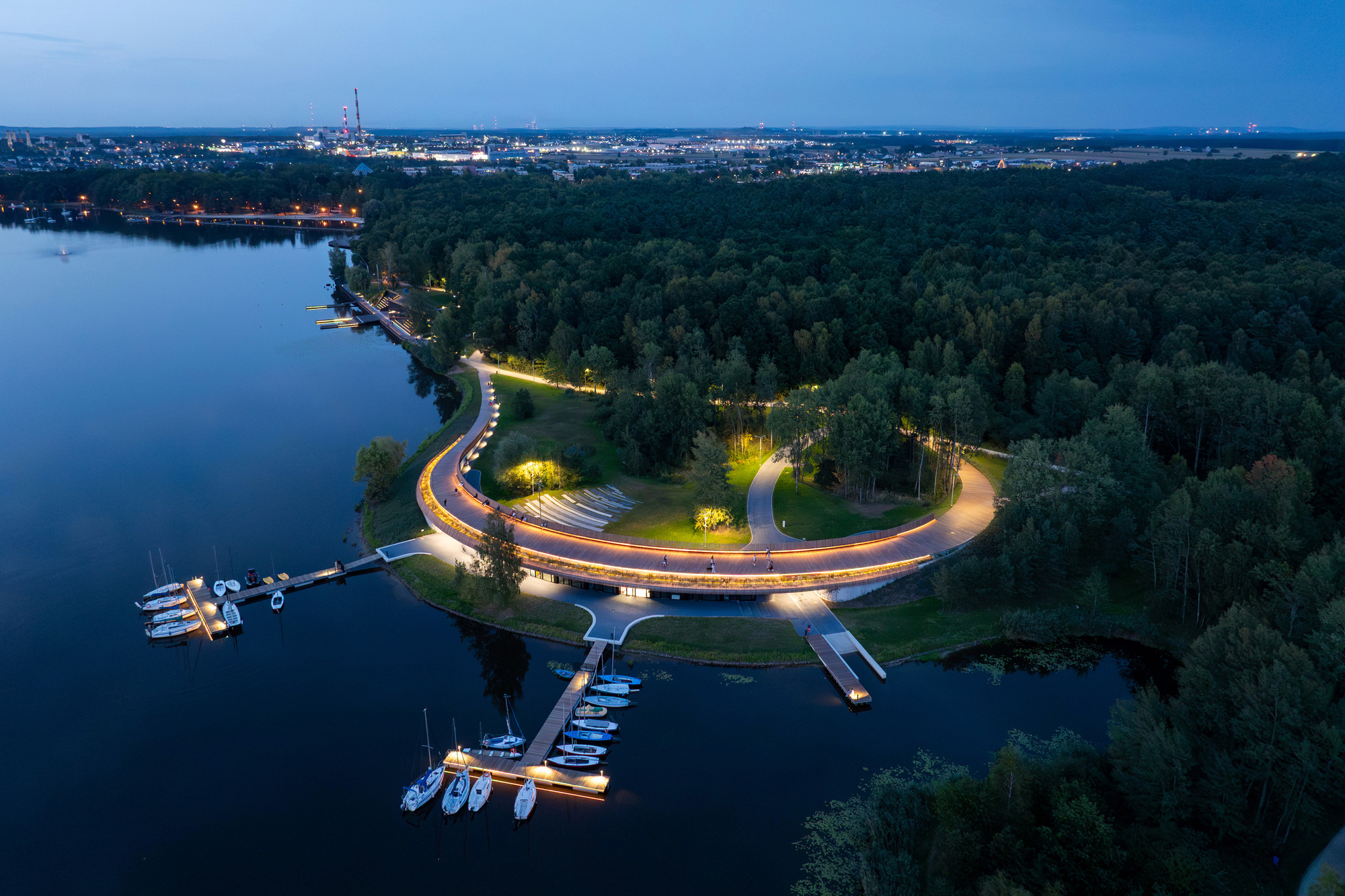 Aerial night view of the glowing curved bridge encircling the wooded peninsula with marina and distant city lights