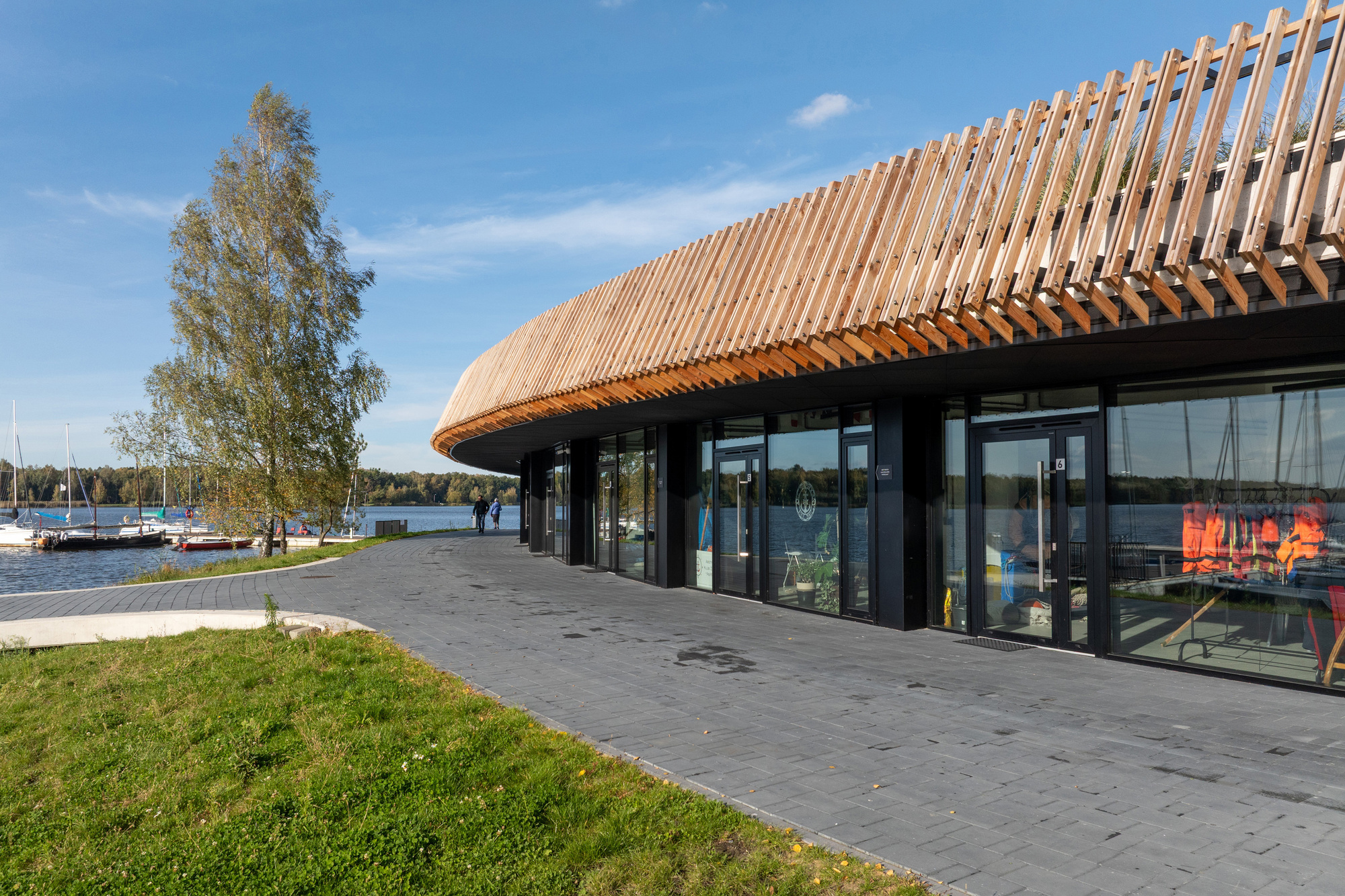 Pavilion facade with full-height glazing and angled timber roof overhang facing the marina under blue sky