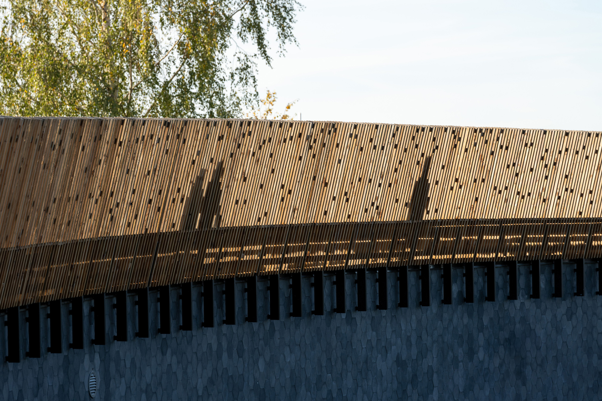Vertical timber screen above a concrete base with shadows of two figures at dusk